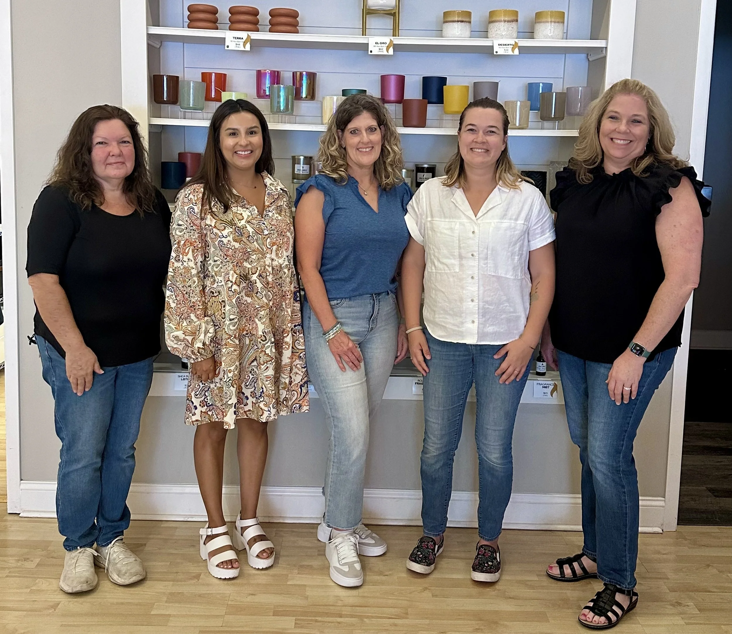 Five women standing side by side indoors in front of a shelf with colorful candles and pottery, smiling at the camera.