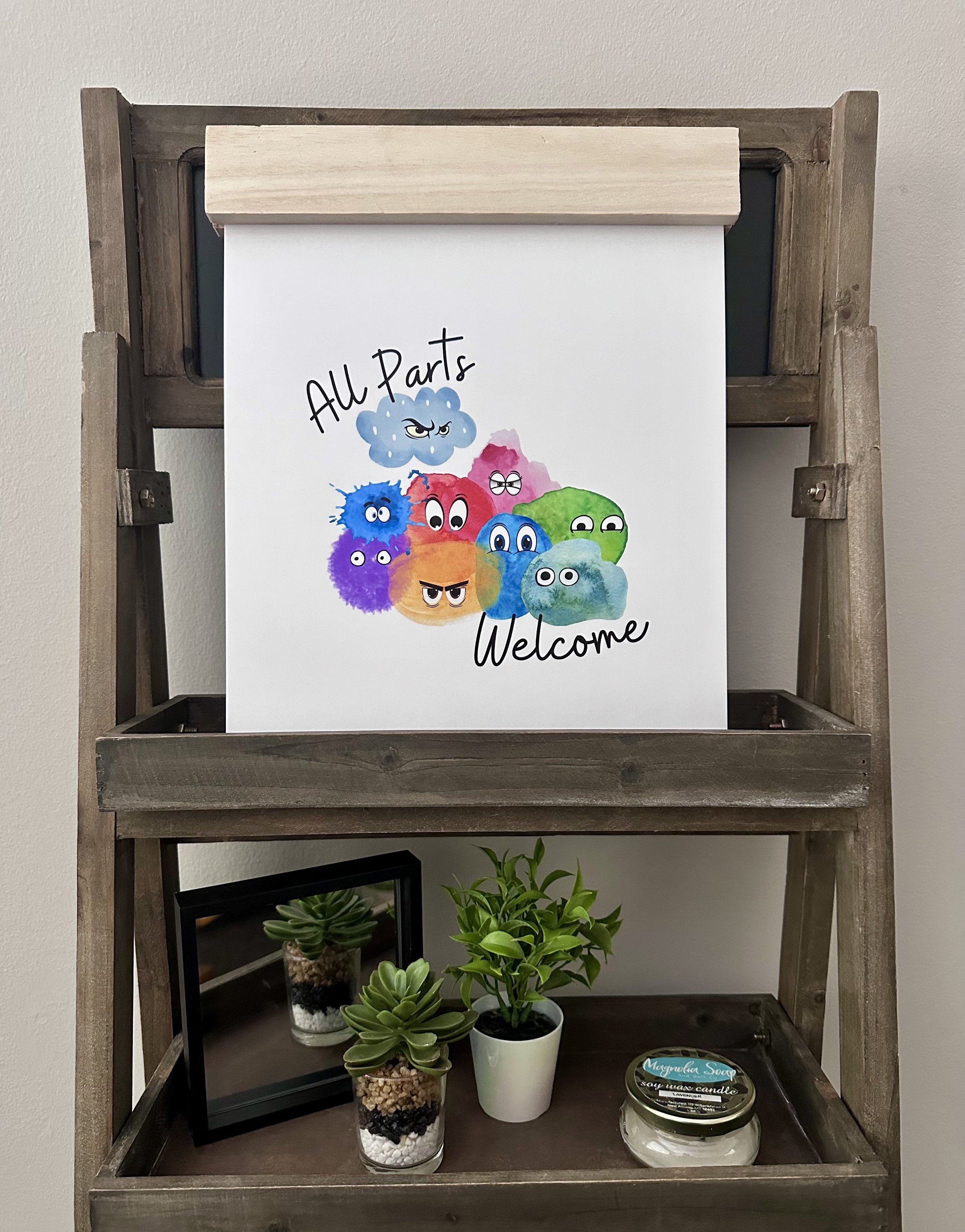 A wooden shelf with a white poster featuring colorful cartoon rocks with faces and the text 'All Parts Welcome.' Below the poster are potted plants and a scented candle.
