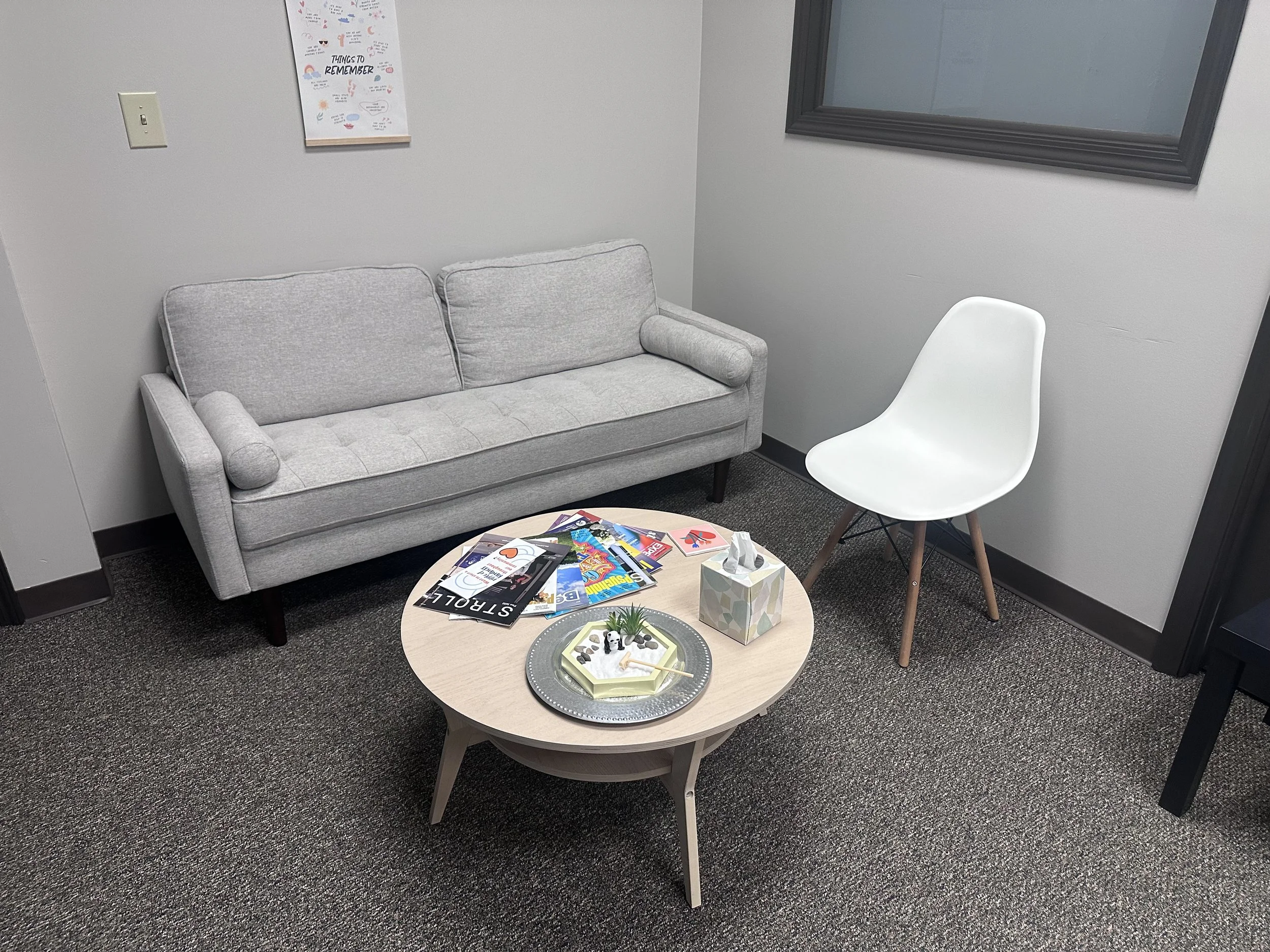 A waiting room with a gray sofa, a white chair, a round wooden coffee table with magazines, a tissue box, a small plant, and decorative stones, with a framed mirror and a colorful motivational poster on the walls.