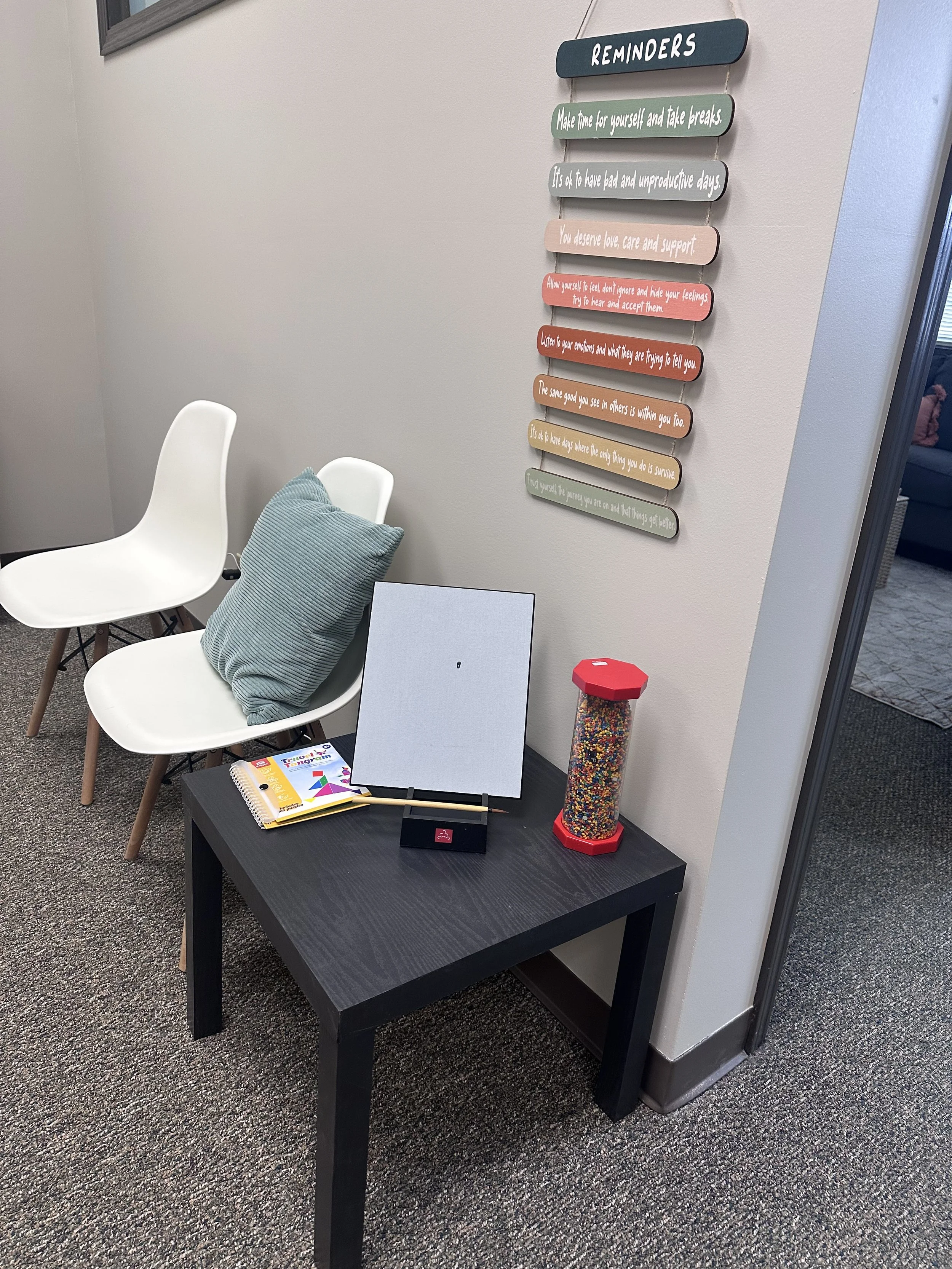 A small black table with a white board and yellow notepad, set against a beige wall with a colorful sign that reads 'Reminders', accompanied by a pen and a container of colorful sprinkles. Nearby, two white chairs, one with a light turquoise pillow. A corner of a room with a gray carpet is visible.
