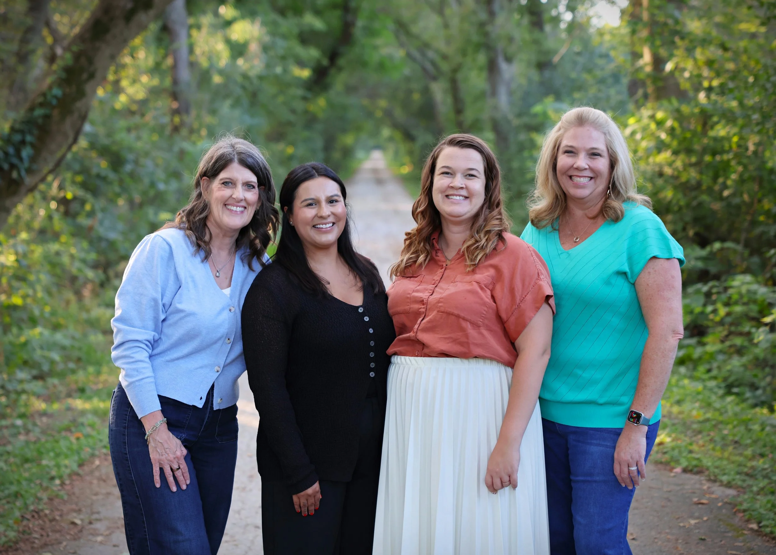 A group of four women standing together outdoors on a dirt path surrounded by green trees, smiling at the camera.