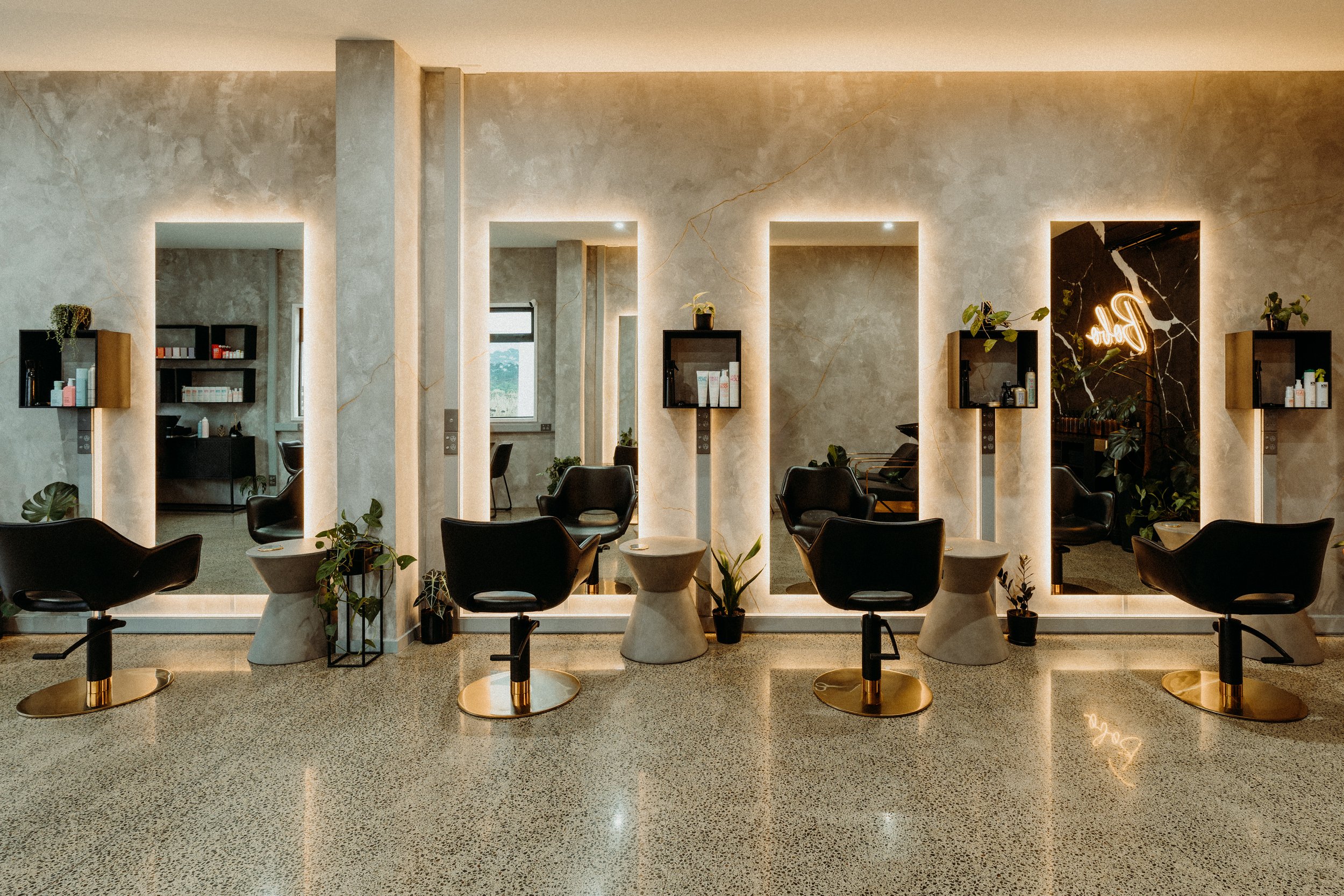 Modern hair salon interior with lighted mirrors, black chairs, and gray marble walls.