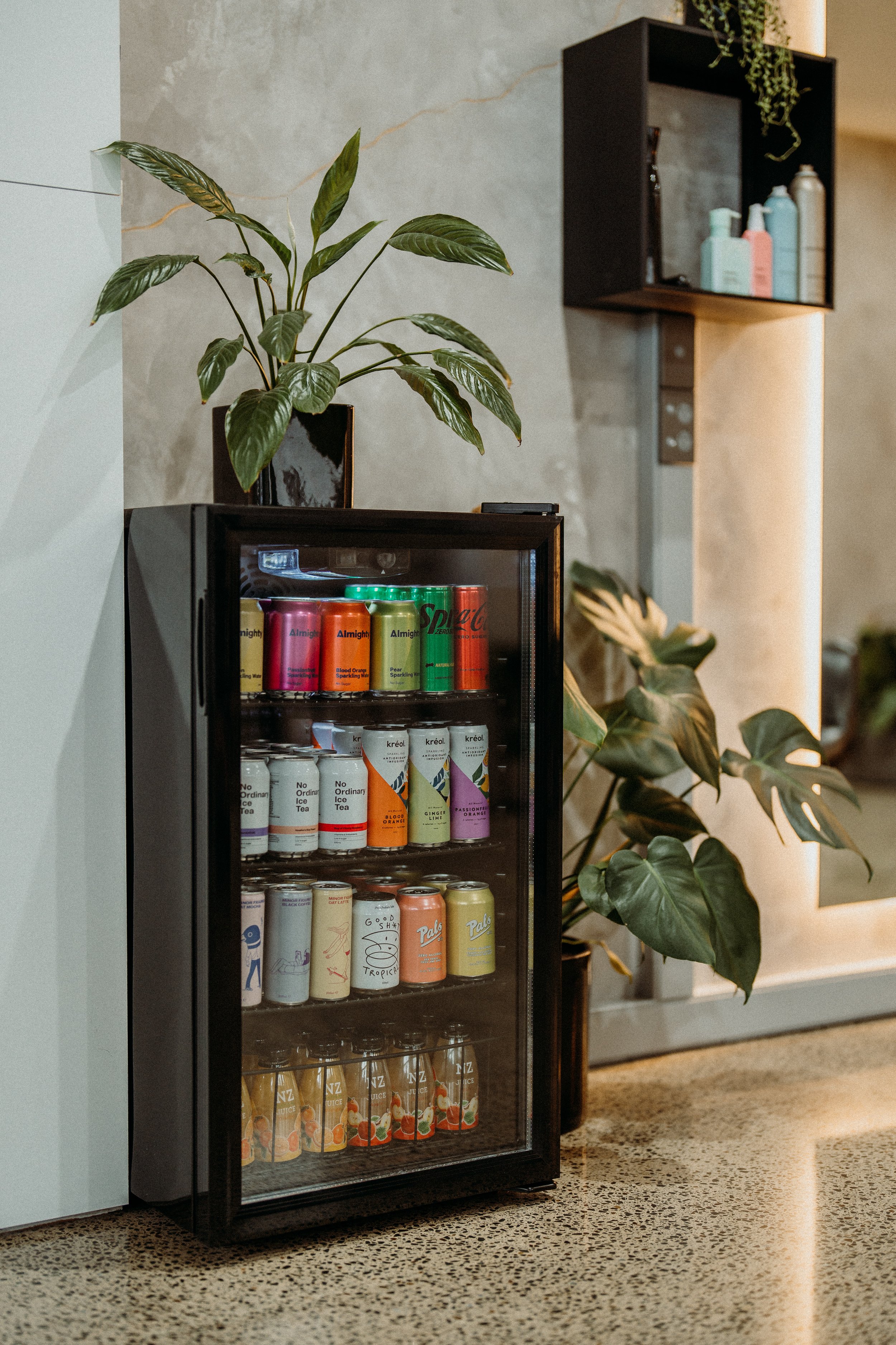 Small beverage fridge with various cans and bottles, plants on top and around, modern interior setting.