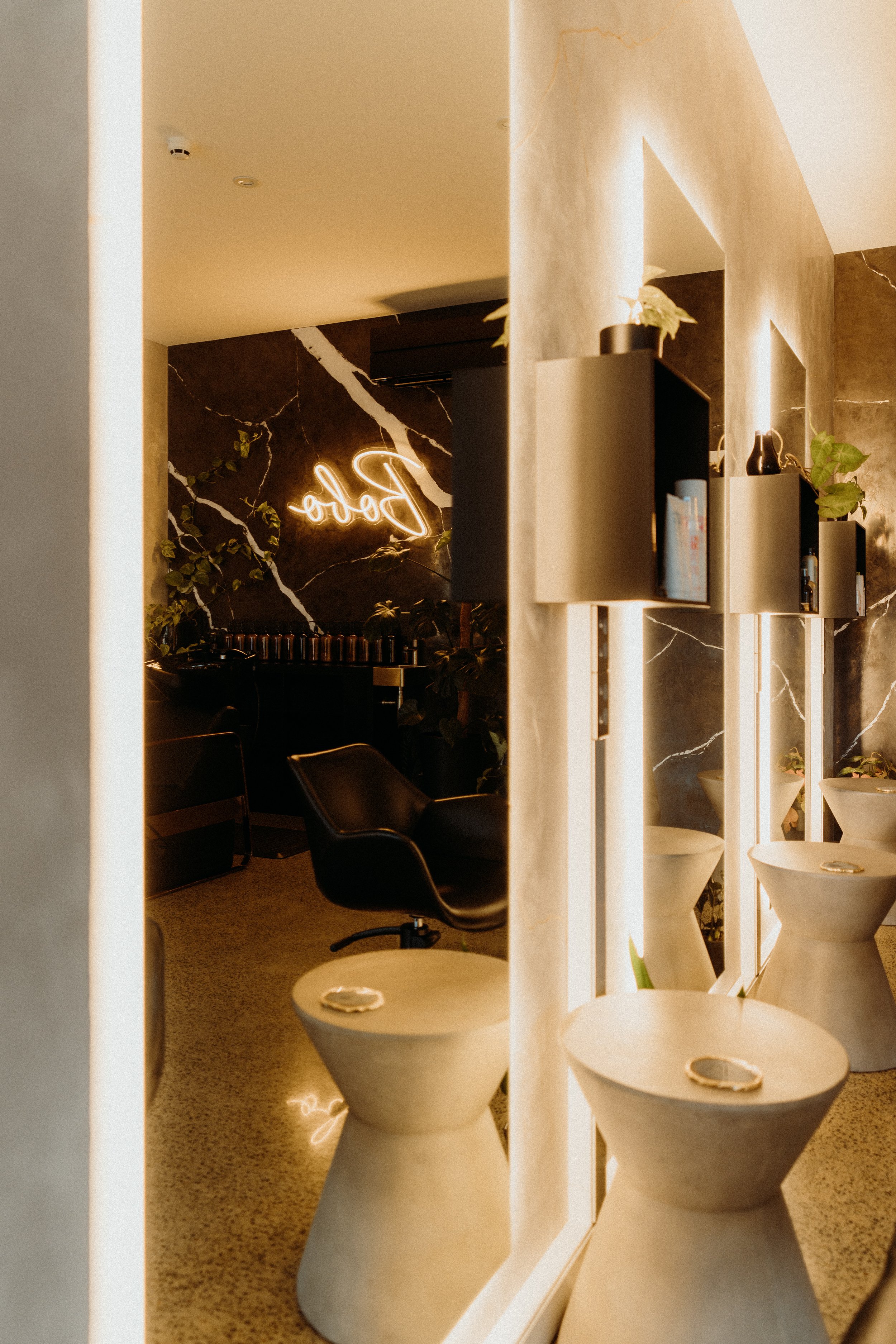 Stylish modern interior with illuminated mirrors, round stools, and a neon "Barber" sign on a marbled wall. Black salon chair and indoor plants add to decor.