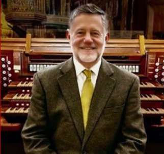 A smiling man in a suit and tie standing in front of a pipe organ.