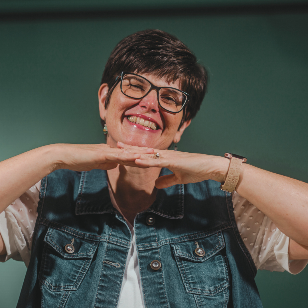 A smiling woman with short dark hair, wearing glasses, a denim vest, and a watch, resting her chin on her hands against a green background.