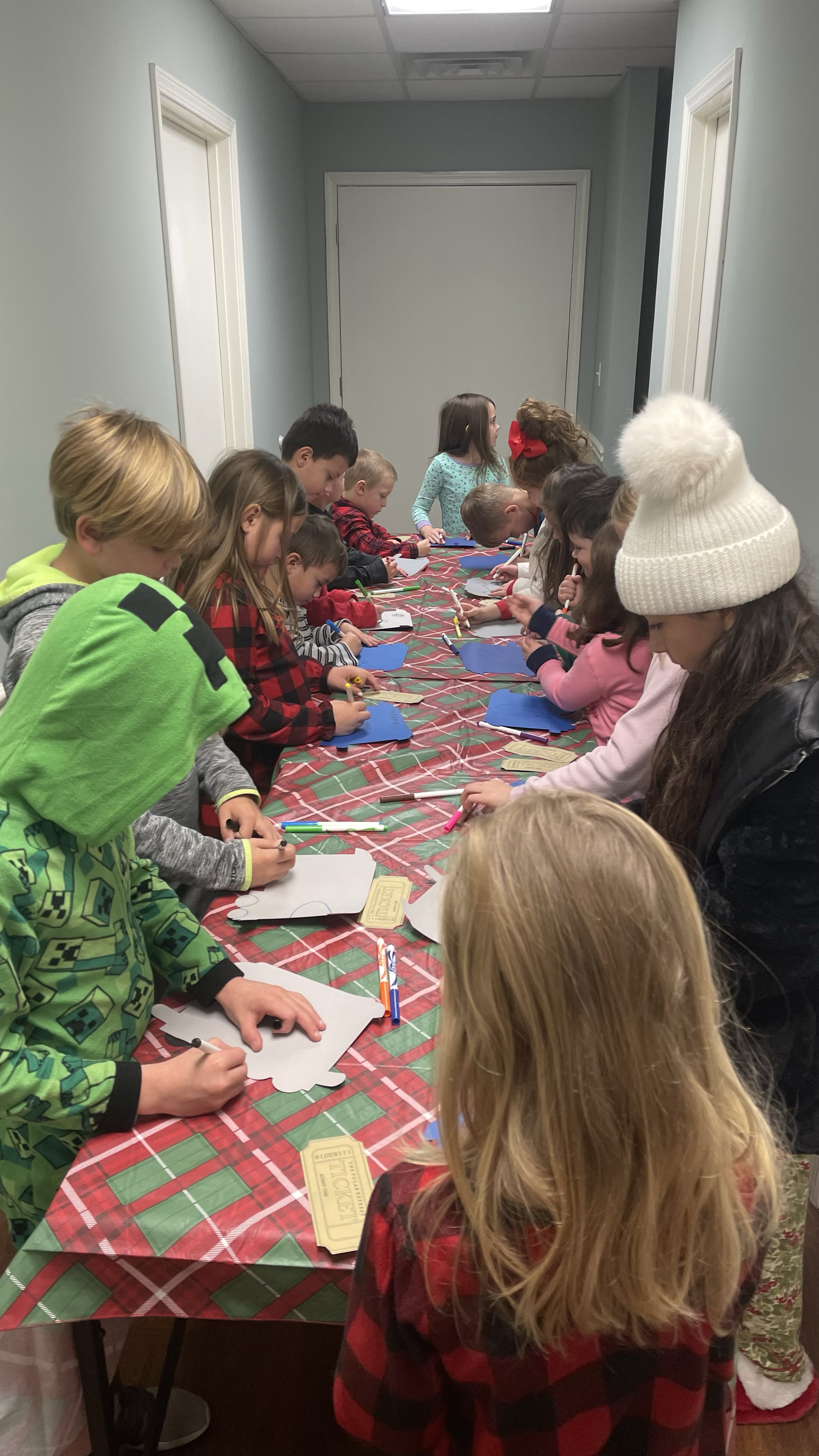 Children gathered around a long table, engaging in holiday craft activities, some wearing festive clothing and accessories.