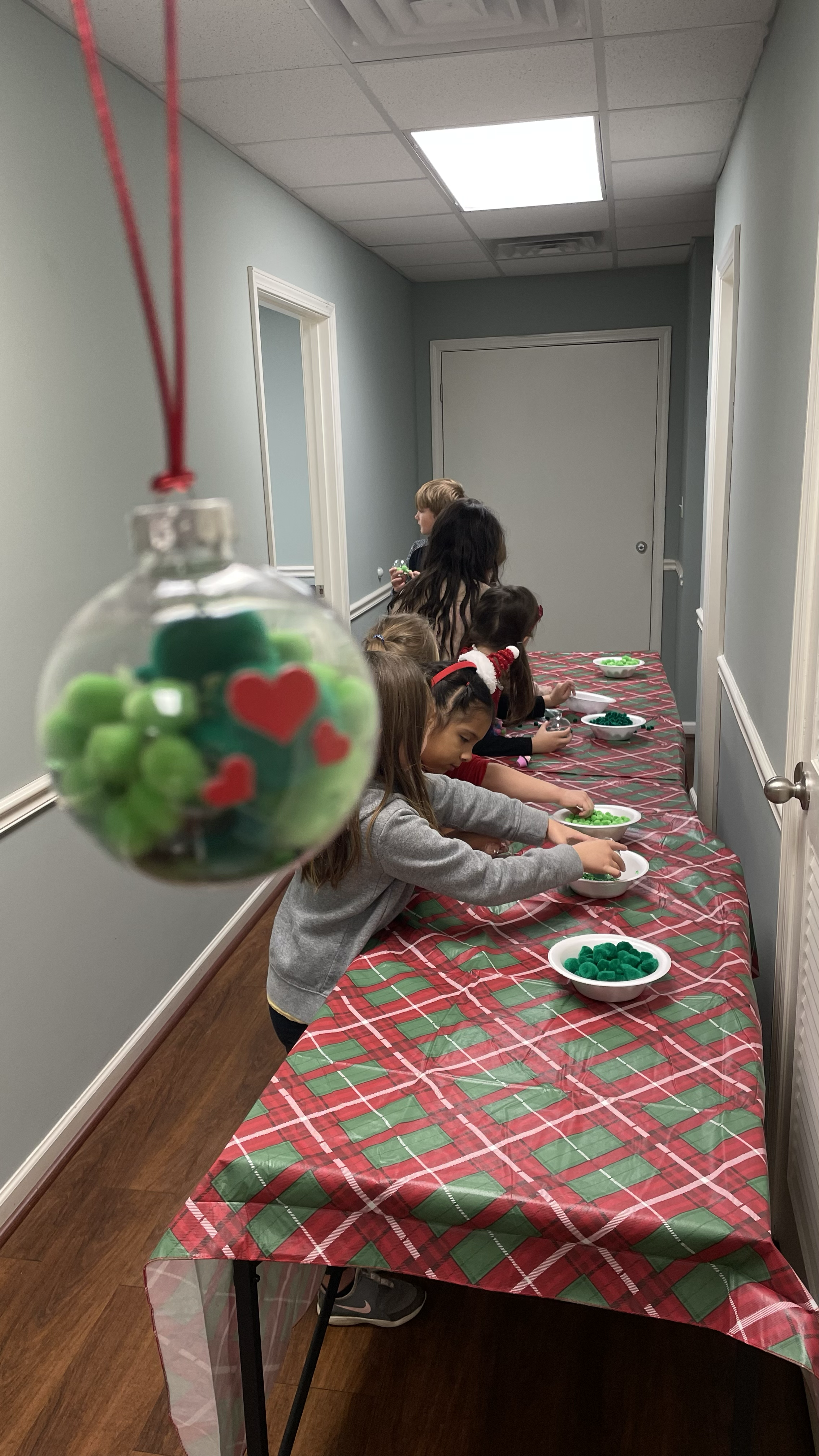 Children participating in a holiday craft activity at a table covered with a red and green plaid tablecloth, with bowls of green craft supplies, in a room with gray walls and closed door.