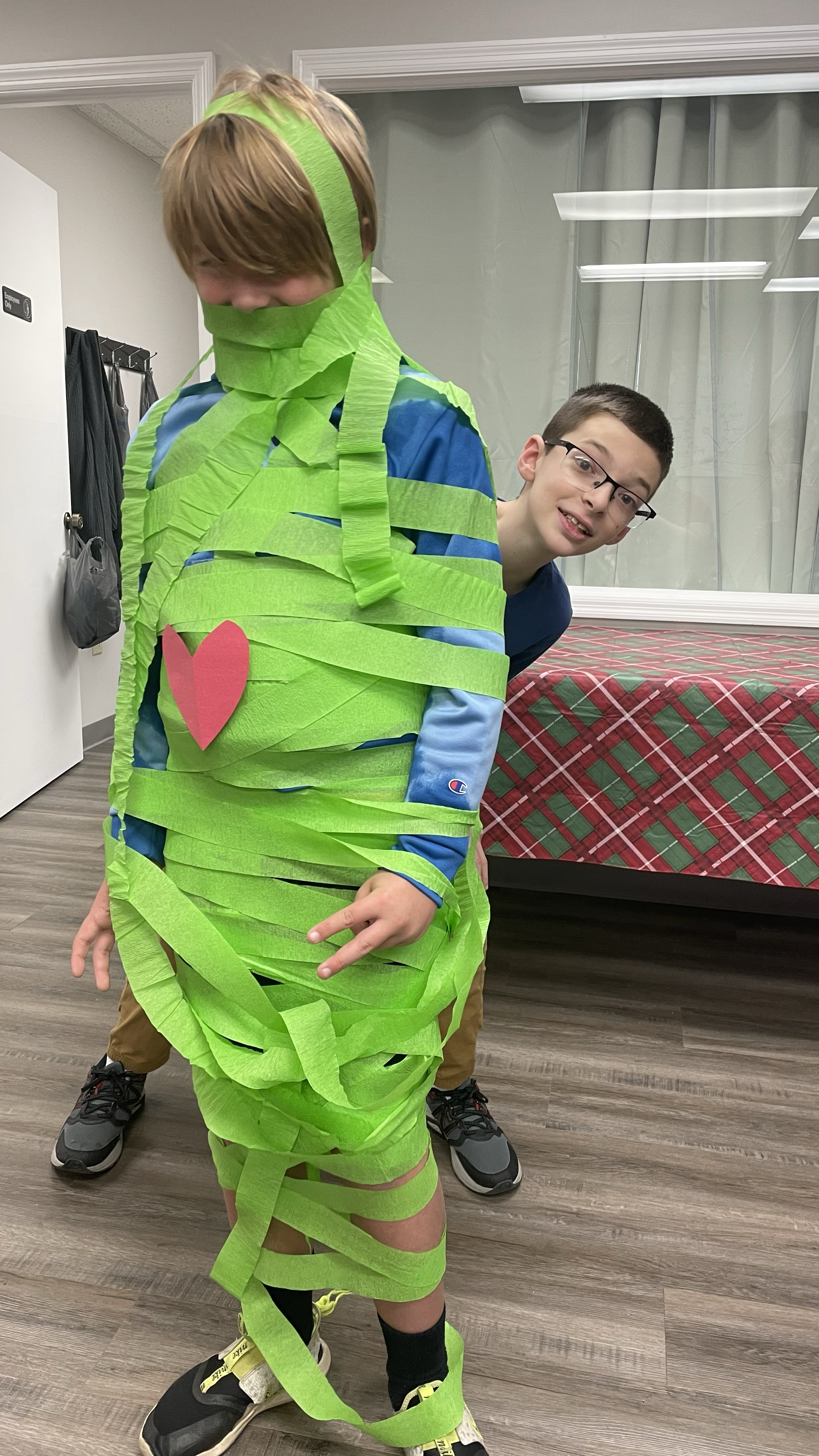 Two boys in a room, one is wrapped in green tissue paper with pink heart decoration, next to a bed with a red and green plaid cover.
