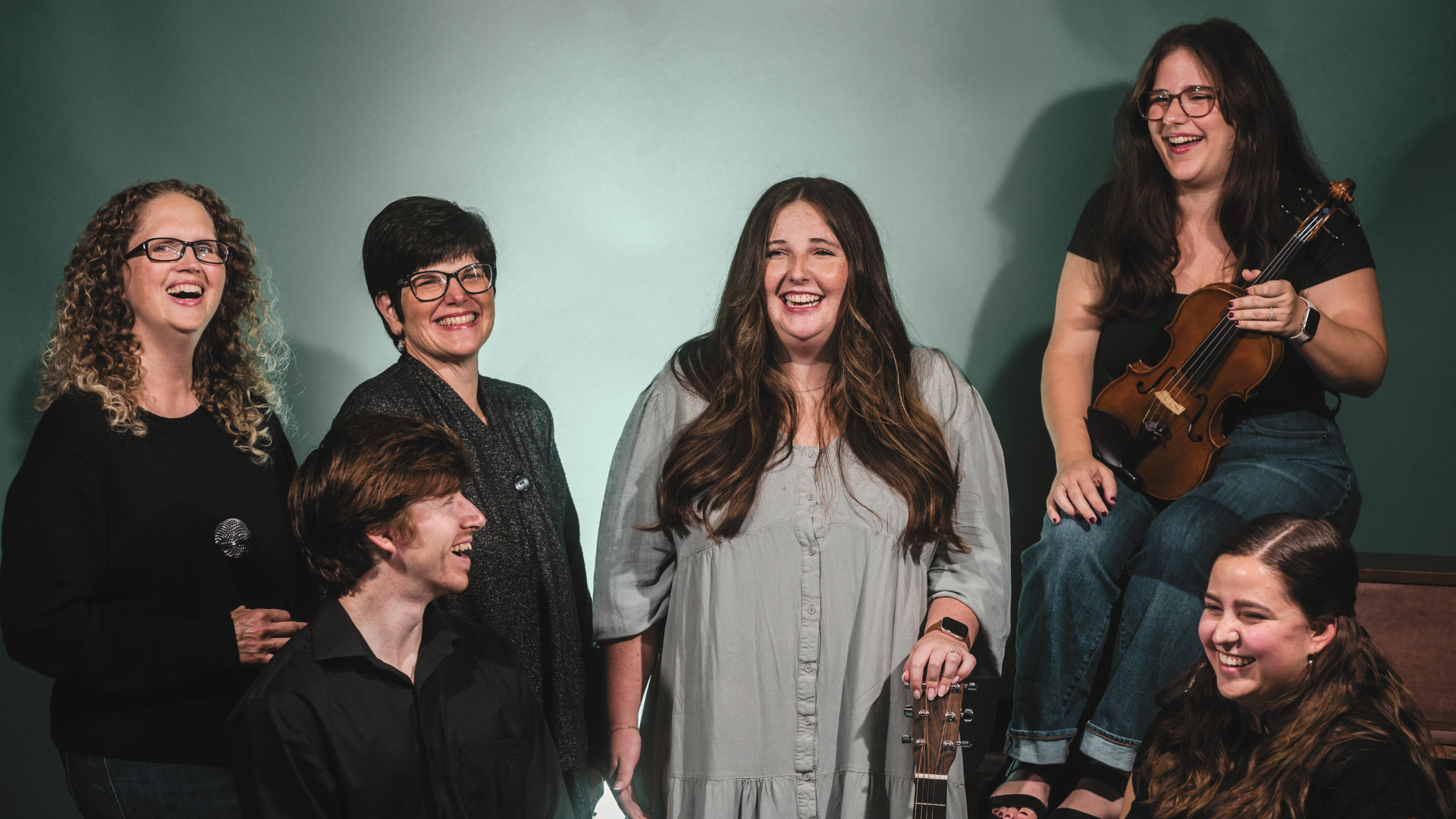 A group of six people, five women and one man, sharing a joyful moment. One woman is sitting on a stool and holding a violin, while the others are standing or sitting nearby, all smiling and laughing.