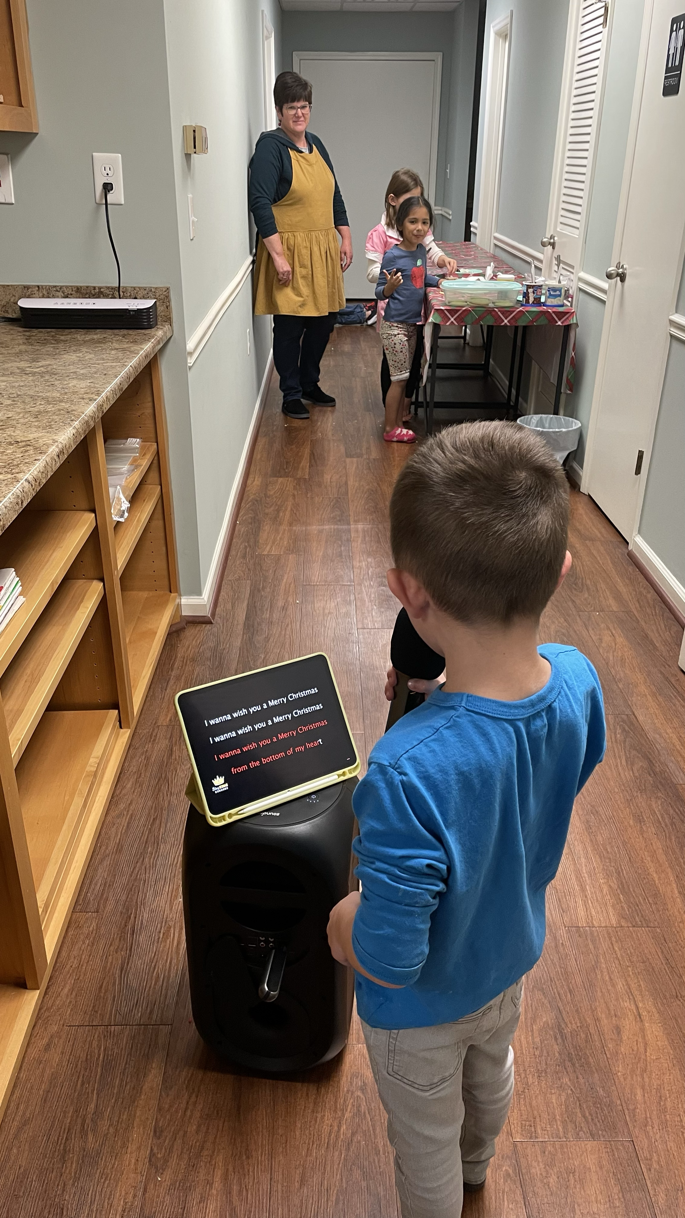 A boy singing karaoke in a hallway, with a tablet displaying Christmas lyrics, while other children and an adult stand near a table in the background.