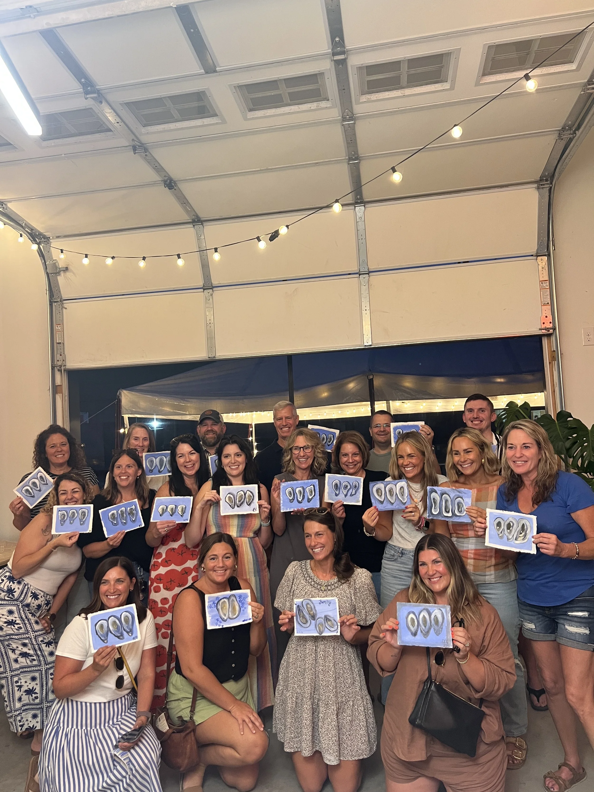 Group of people posing with artwork in a garage turned art space, illuminated by string lights.