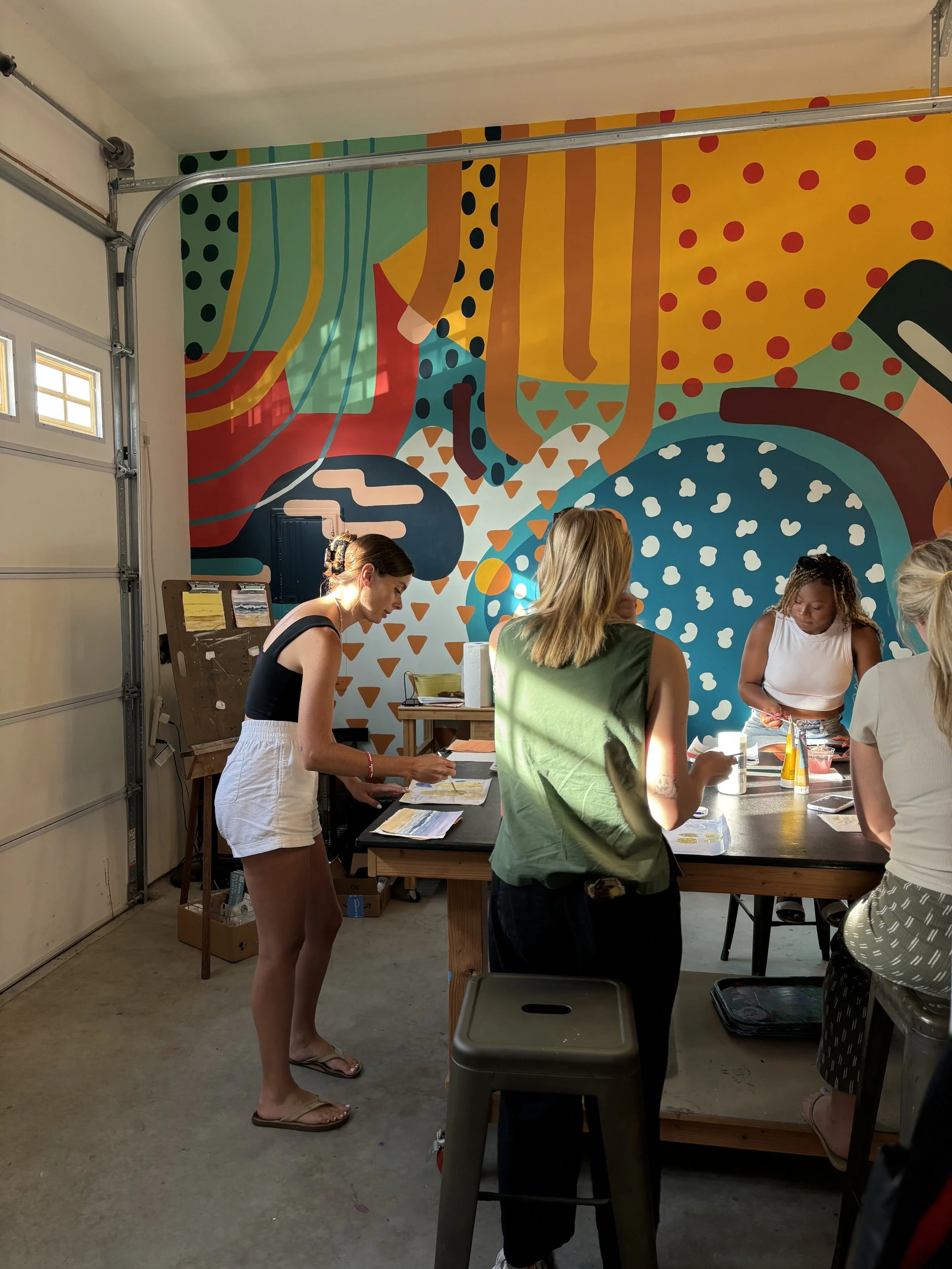 Women gathered around a table in front of a colorful, abstract mural in a garage space.