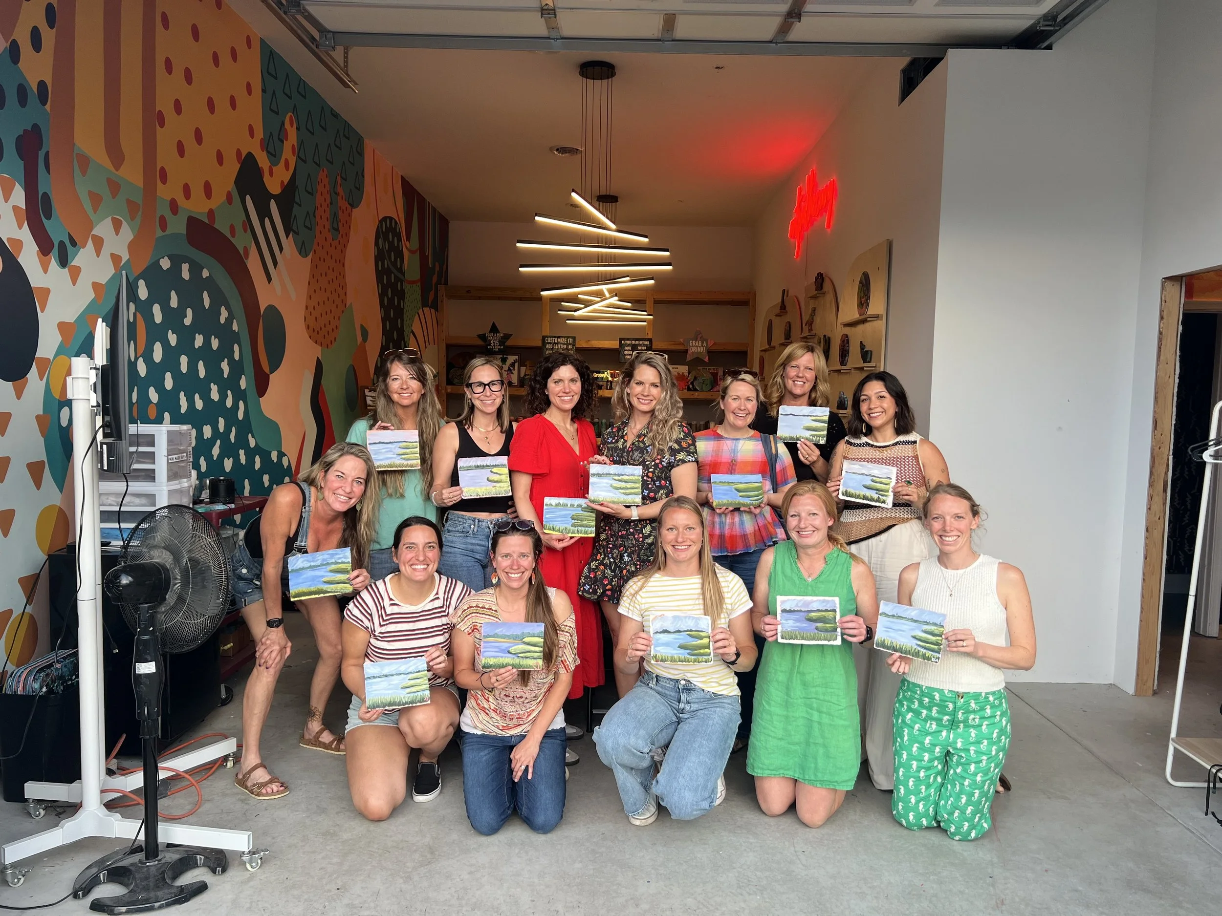 Group of fifteen women in colorful casual clothing, some kneeling and some standing, holding small landscape paintings in a room with a vibrant abstract mural wall and modern ceiling lights, celebrating a painting event.