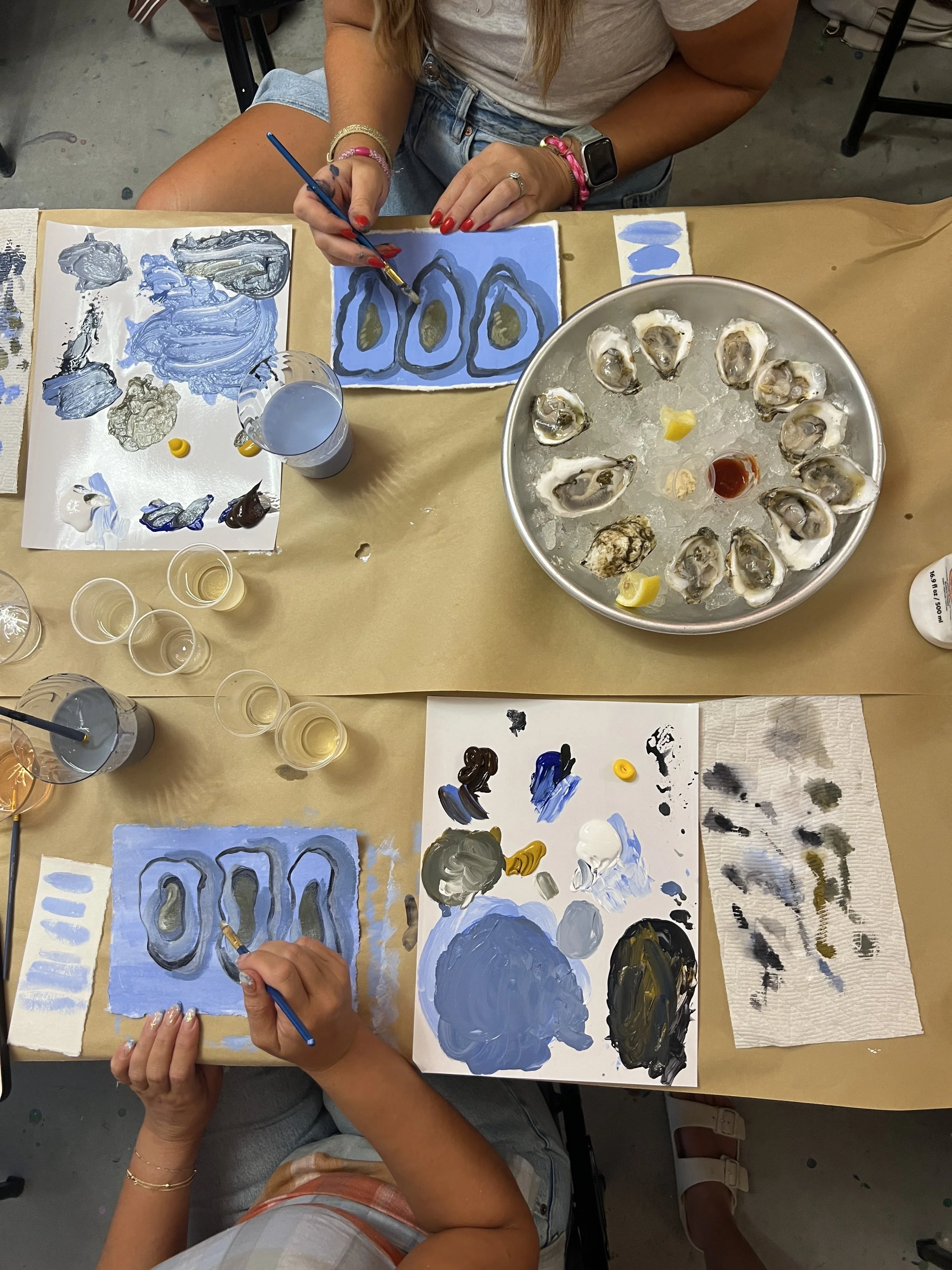 Two people painting sea-themed artwork at a table with oyster shells and paint supplies.