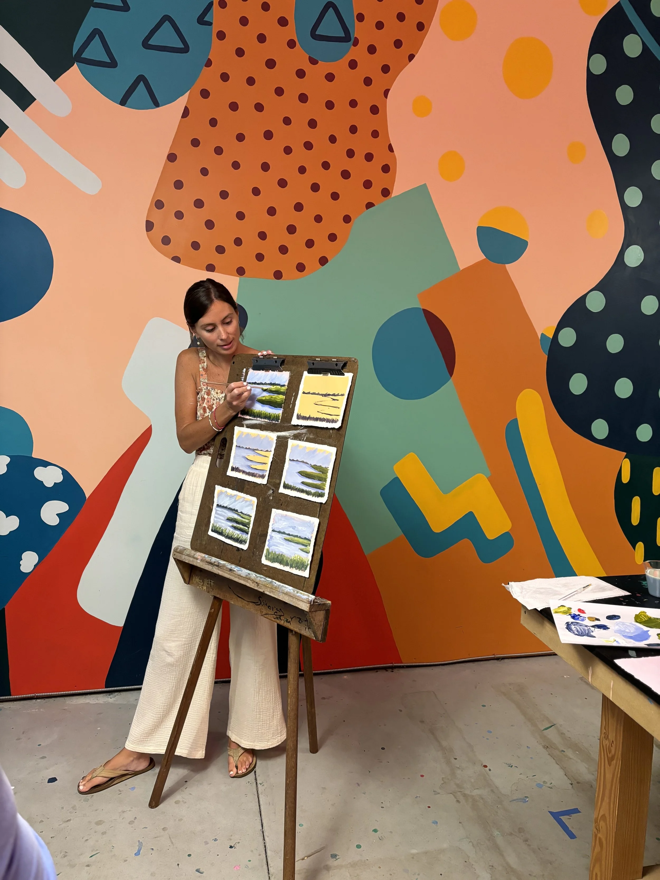 A woman painting landscape scenes on small canvases in front of a large, colorful abstract mural wall.