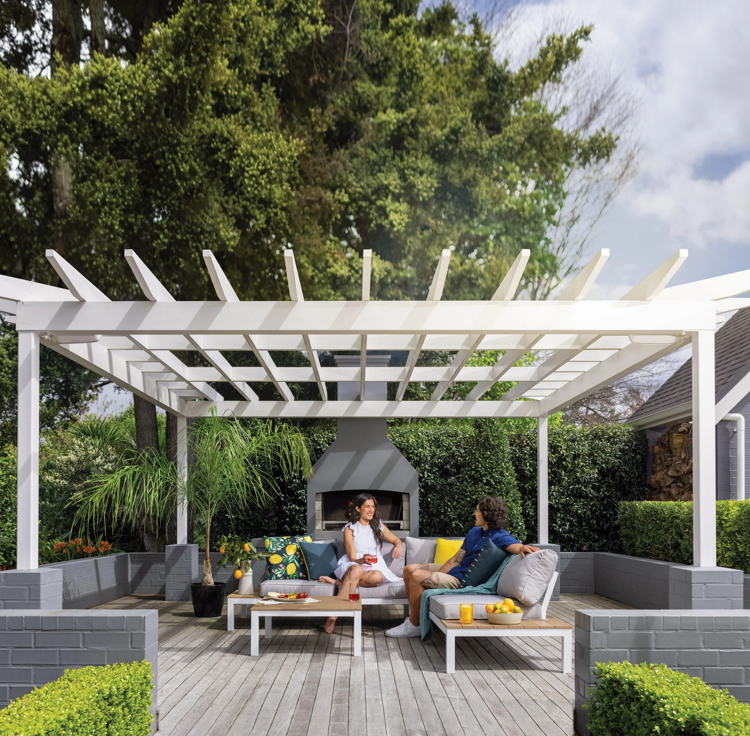 Two people sitting and talking in a backyard patio with a white pergola, gray brick walls, a fireplace, and greenery. An advertisement for a homewares store.