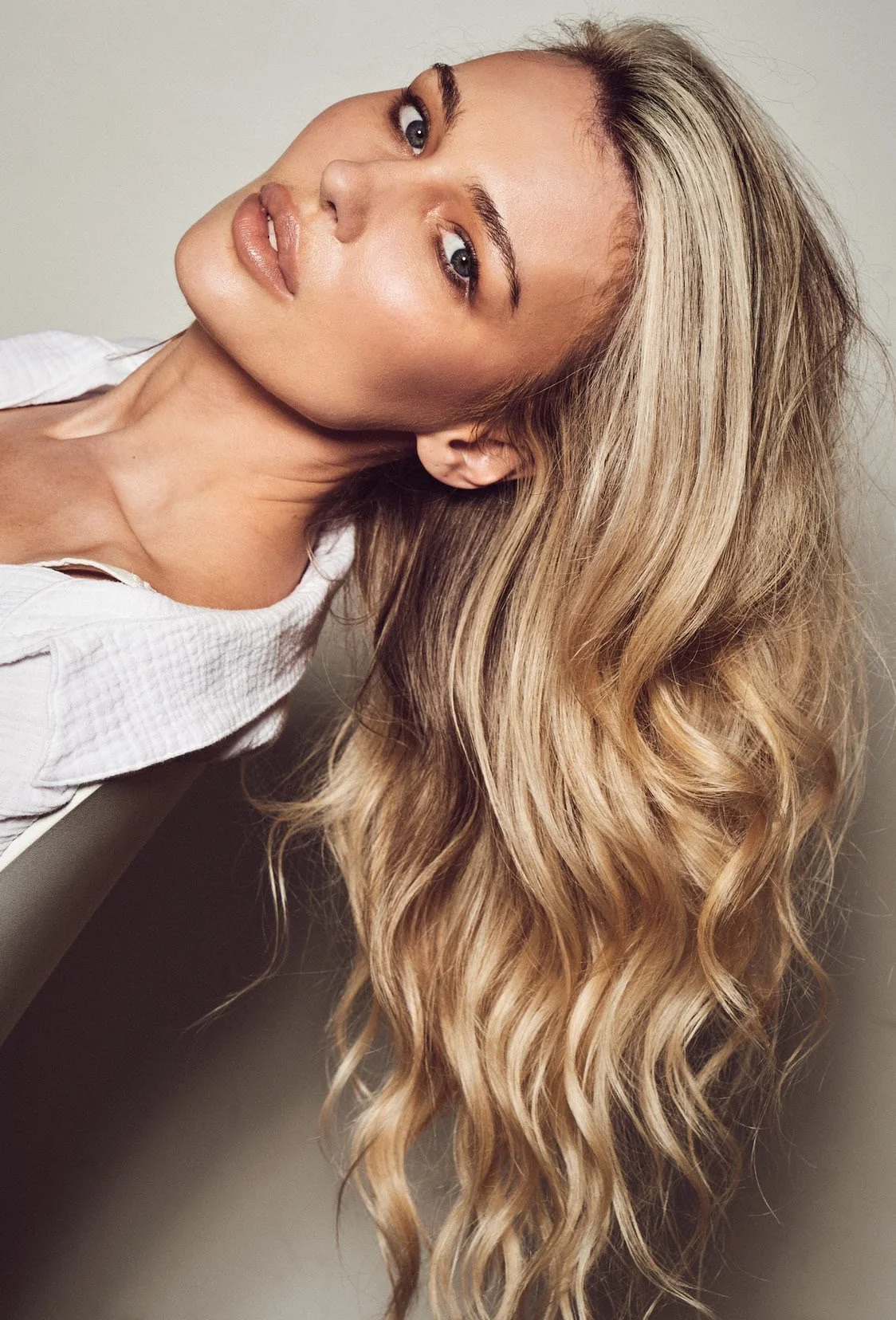 A woman with long, wavy blonde hair tilting her head back slightly, wearing a white shirt.