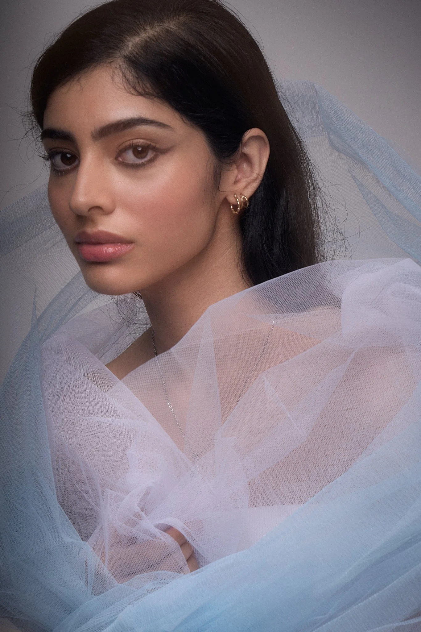 Close-up portrait of a young woman with dark hair, wearing gold earrings and sheer white fabric draped around her shoulders.