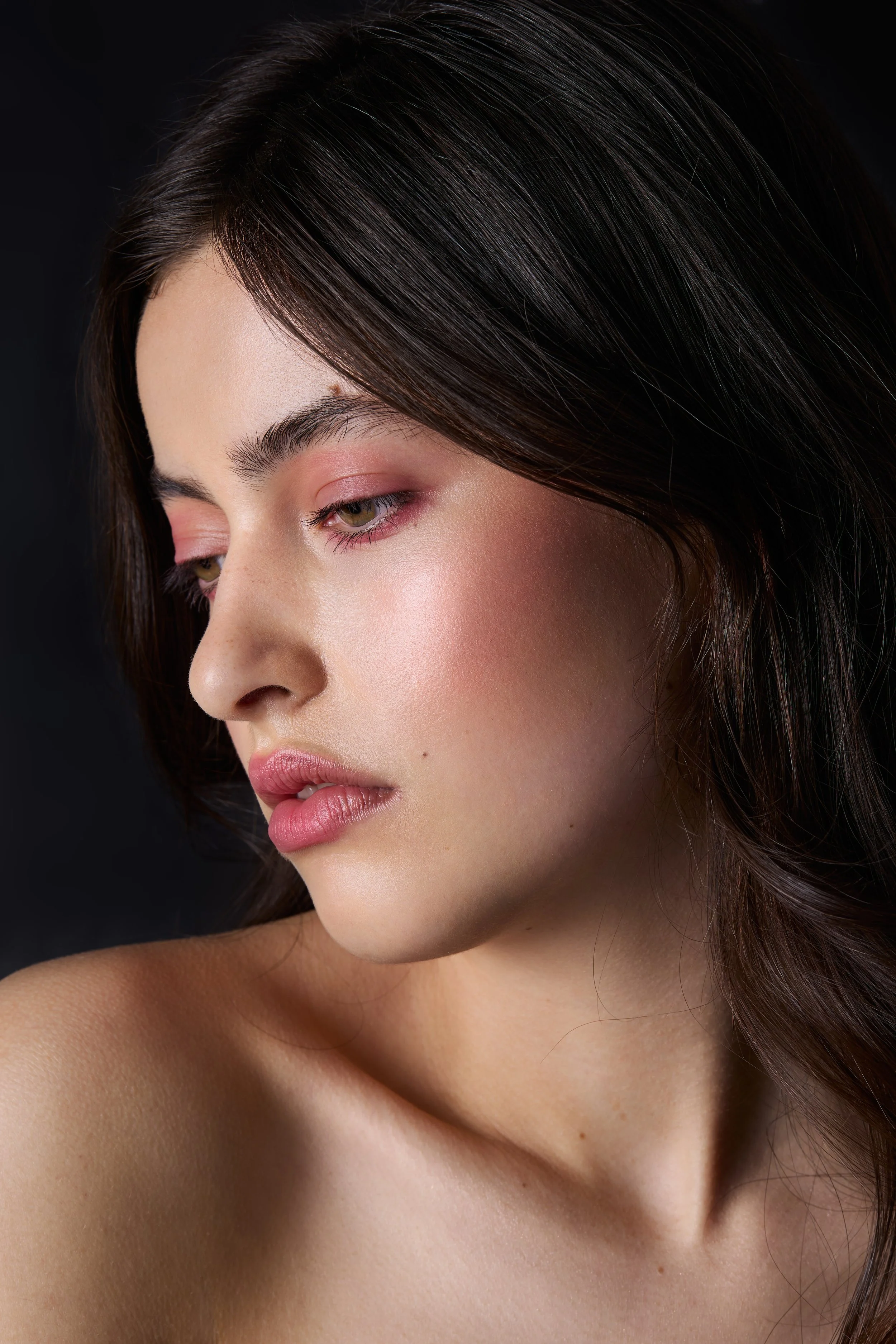 Close-up portrait of a young woman with dark brown hair, soft makeup, light pink eyeshadow, and pink lipstick, looking down against a dark background.