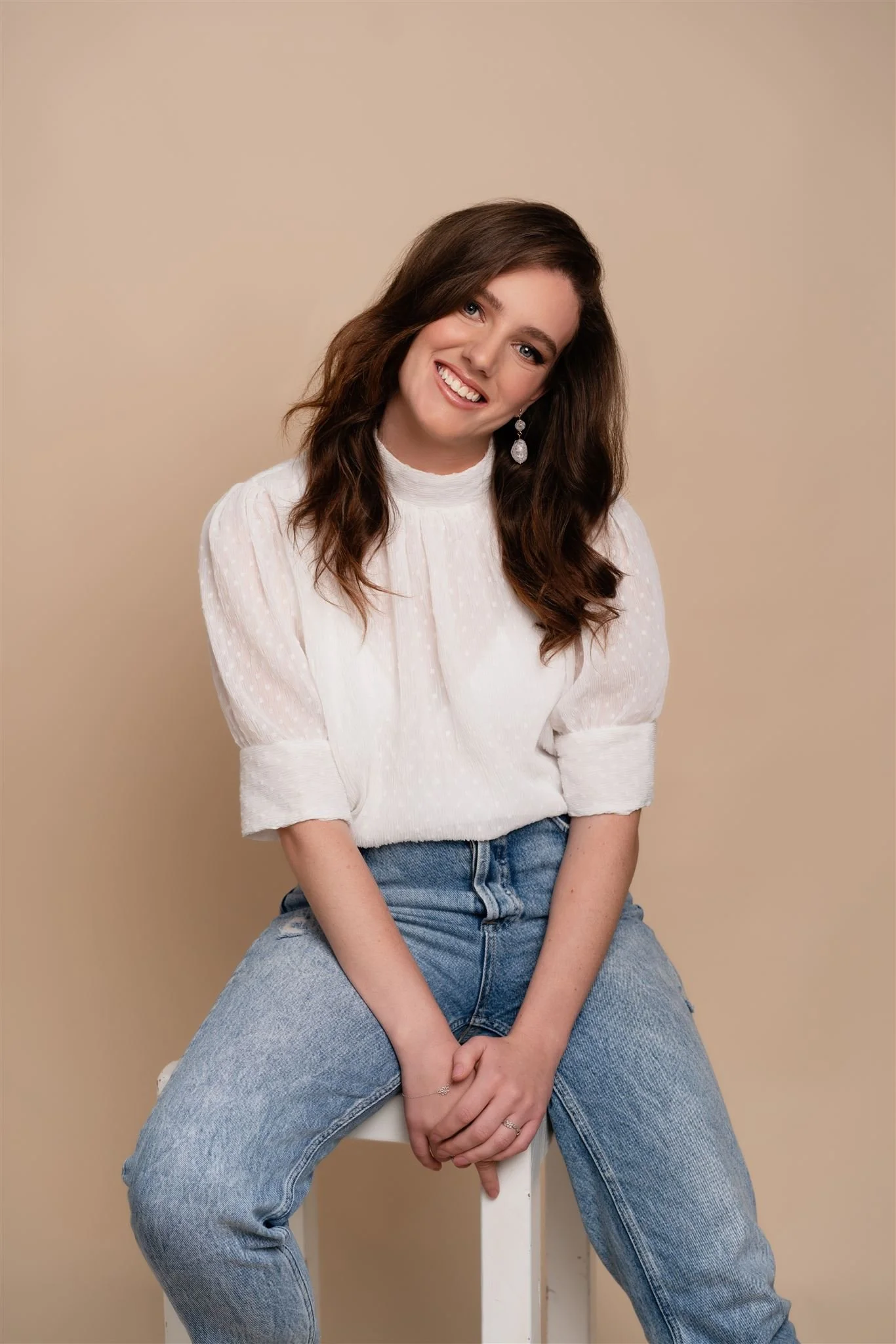 A young woman with wavy brown hair sitting on a white stool against a beige background, smiling and wearing a white blouse with puffed sleeves and large earrings.