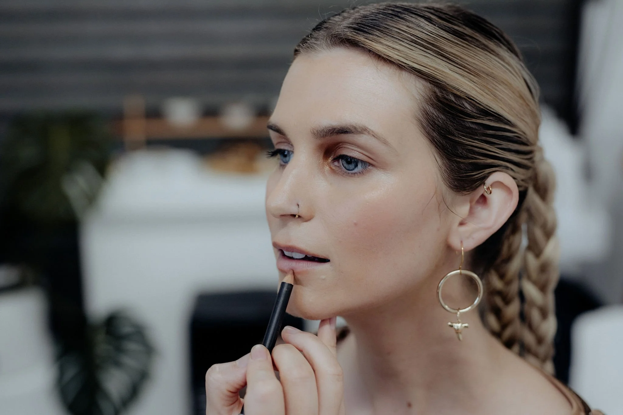A woman with blonde hair in braids is applying lip liner with a pencil, wearing gold hoop earrings with charms, in a modern indoor setting.