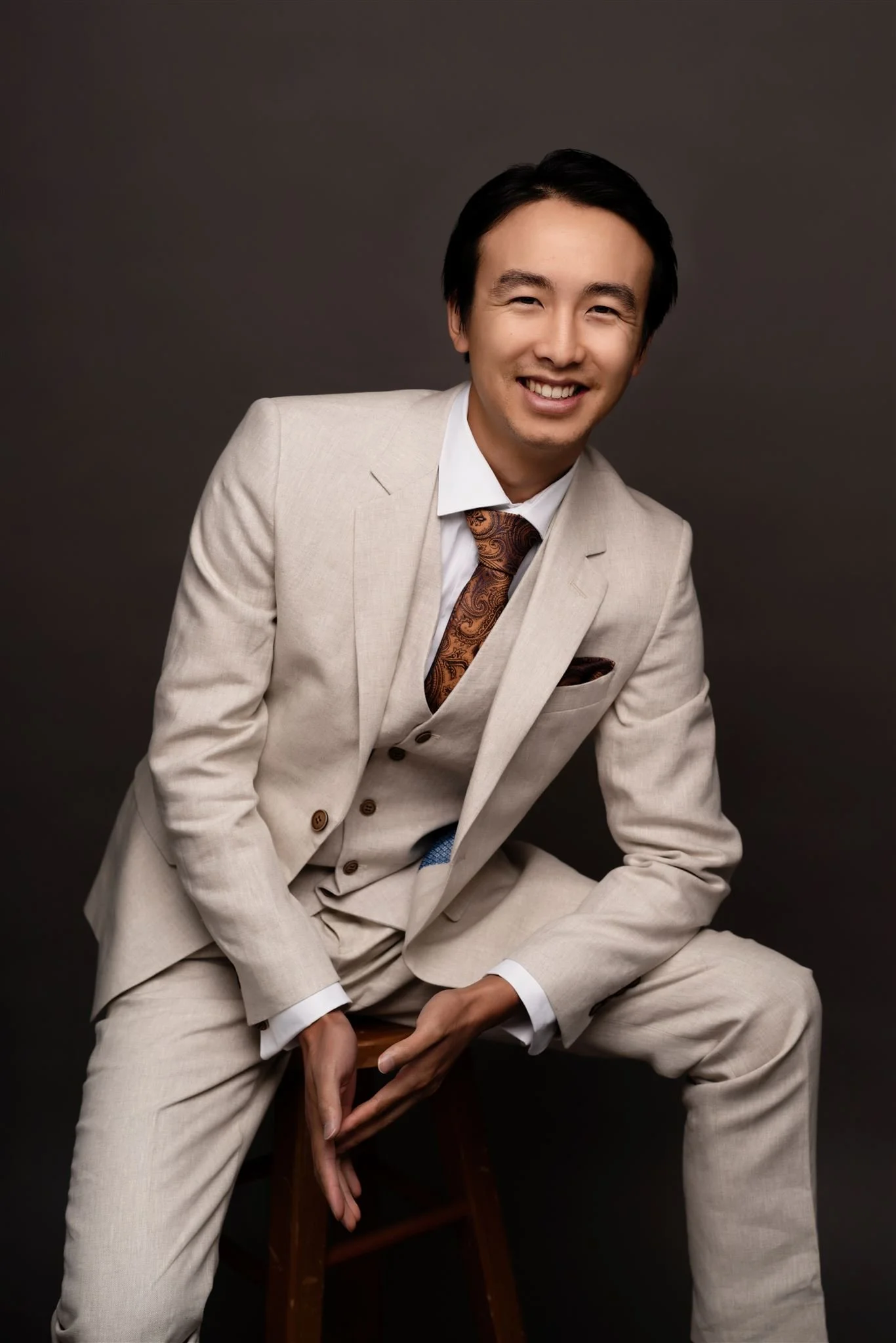 Man in a beige suit, white shirt, and paisley tie, sitting on a stool, smiling against a dark background.