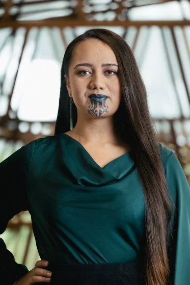 A woman with long dark hair in a teal top, wearing cyberpunk-style makeup and a futuristic mask covering her mouth, standing indoors with large windows and wooden beams in the background.