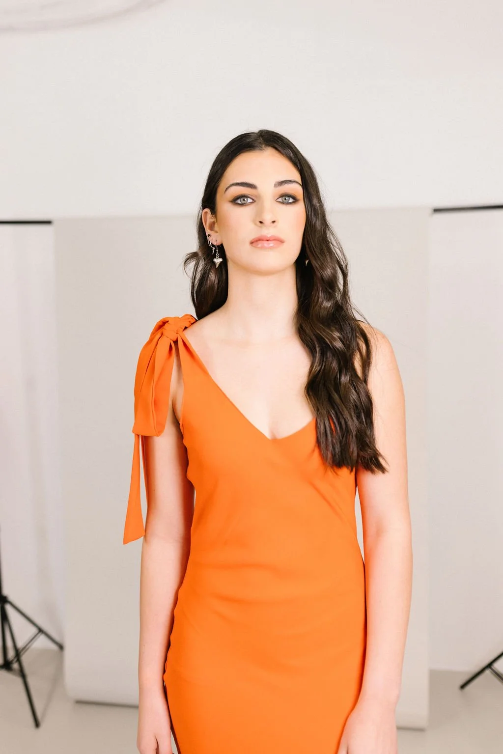 A woman with long dark wavy hair and light skin wearing an orange dress with a bow on the shoulder, posing in front of a neutral background.