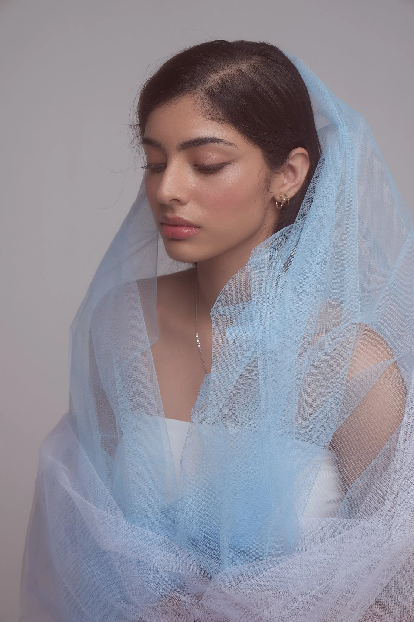 A woman with dark brown hair, closed eyes, and gold hoop earrings, wrapped in light blue sheer fabric, light makeup on a neutral background.