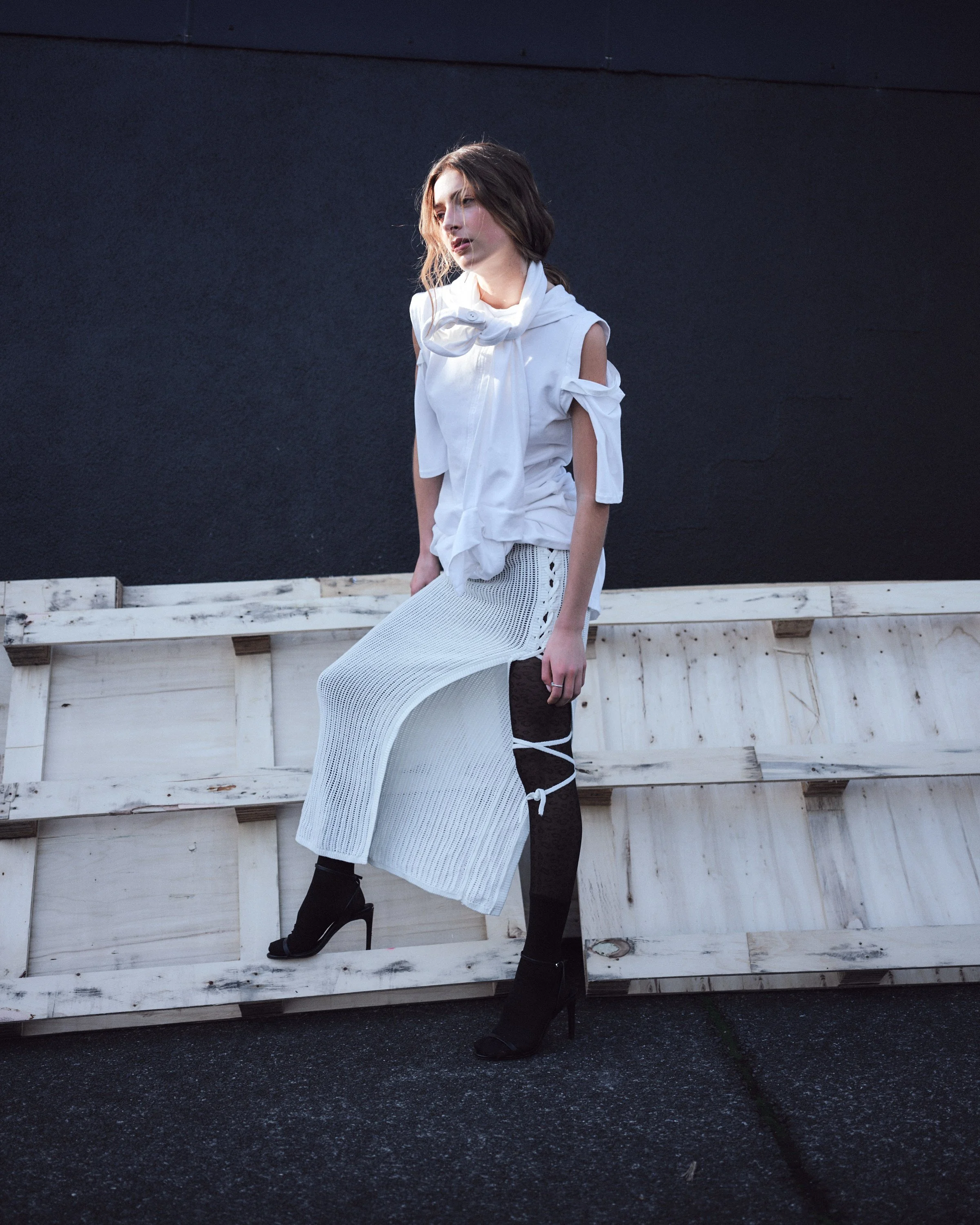 A woman wearing a white top with cut-out shoulders, a white knee-length knitted skirt, and black high-heeled boots stands against a dark wall and a wooden barrier.