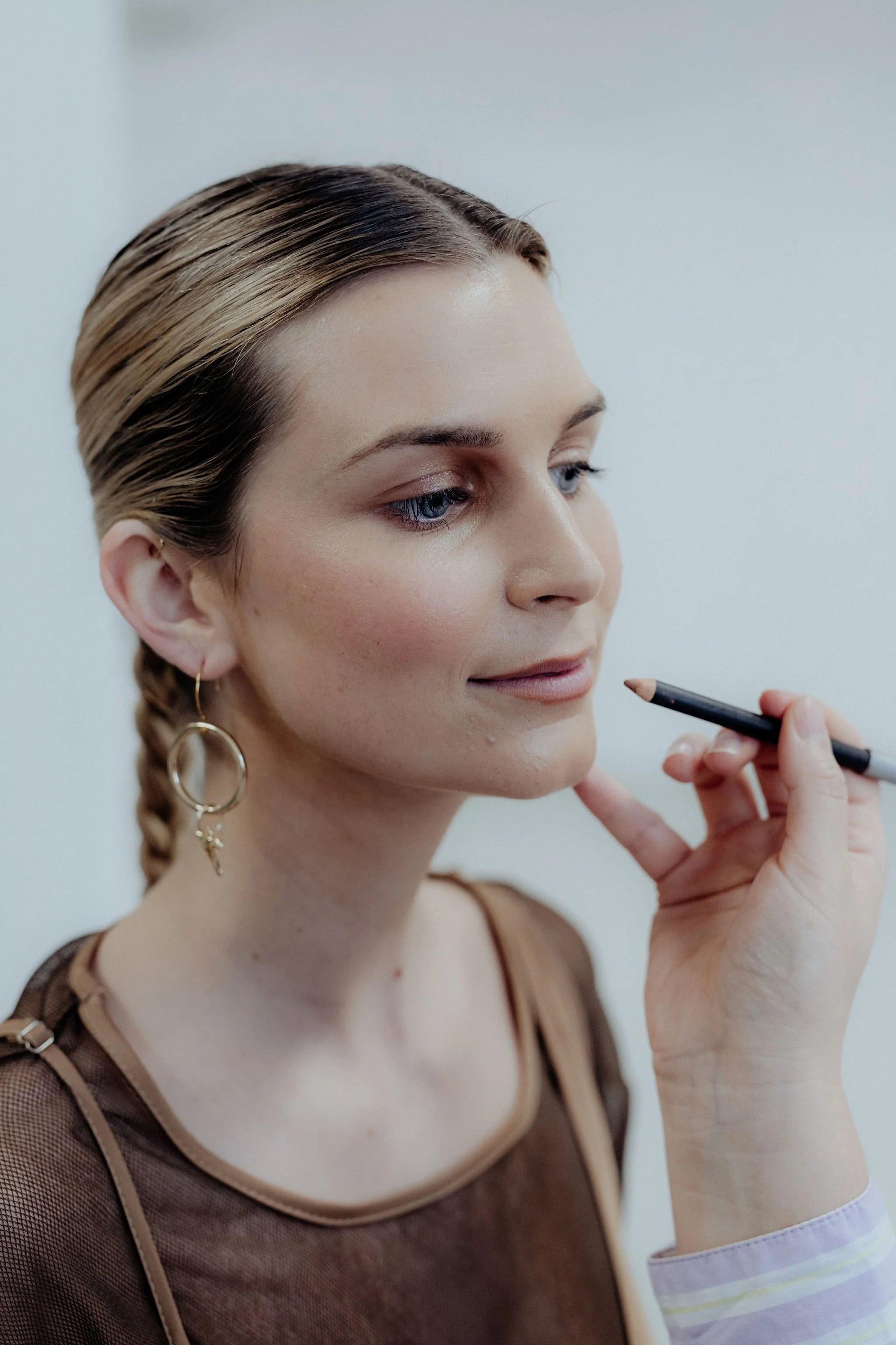 A woman with braided hair and earrings is having makeup applied to her lips with a pencil by a makeup artist.