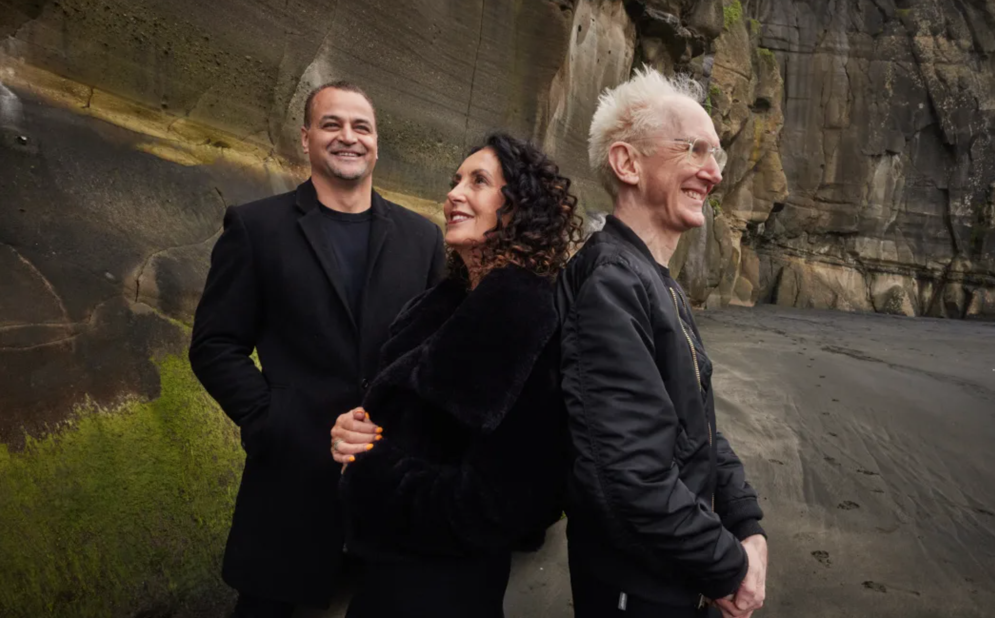 Three people standing outdoors near a rocky cliff, smiling and looking in different directions.