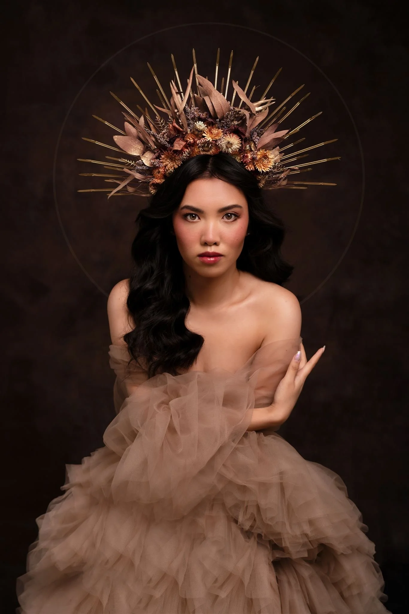A woman with long dark wavy hair wears a beige, tulle dress and a floral headpiece with gold accents, set against a dark background.