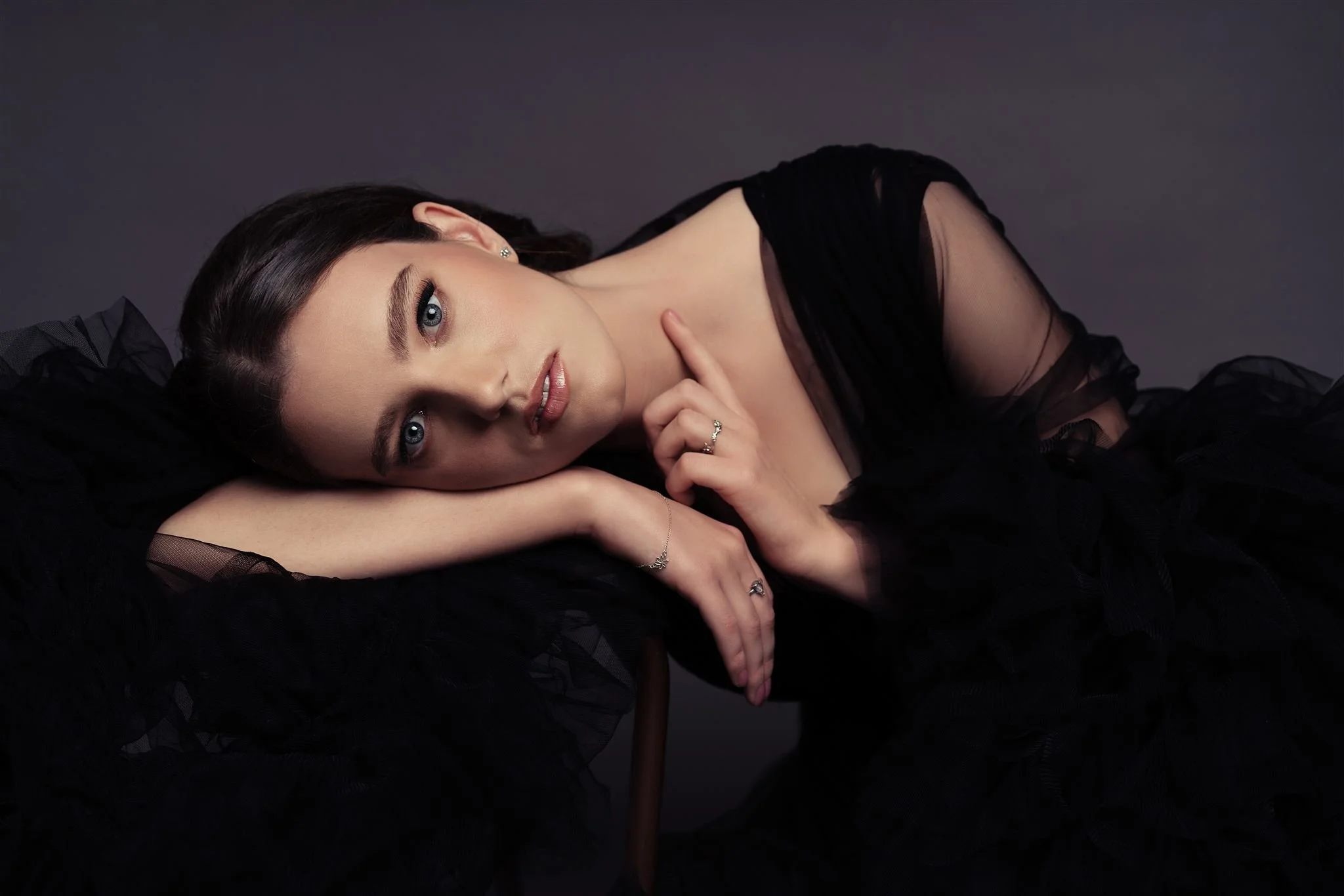 A young woman with blue eyes and dark brown hair, wearing a black sheer dress, lying down with her head resting on her arm, looking at the camera against a dark background.