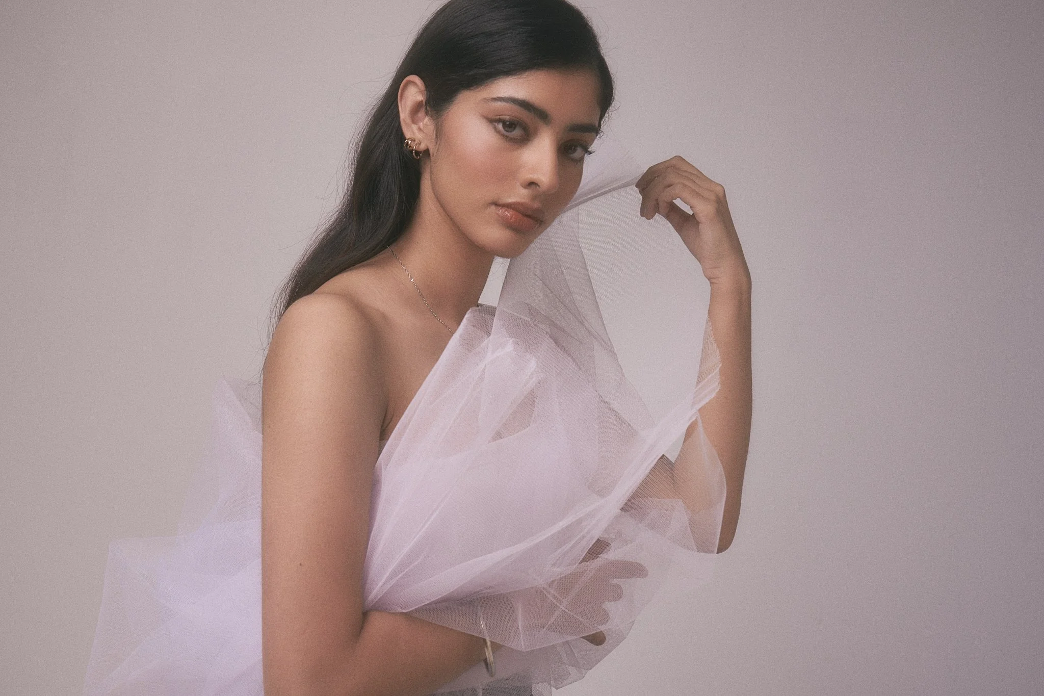 A young woman with long dark hair, wearing gold hoop earrings and a delicate necklace, poses against a plain light background. She is partially wrapped in white tulle fabric, holding some of it near her face and shoulder, with a neutral expression.