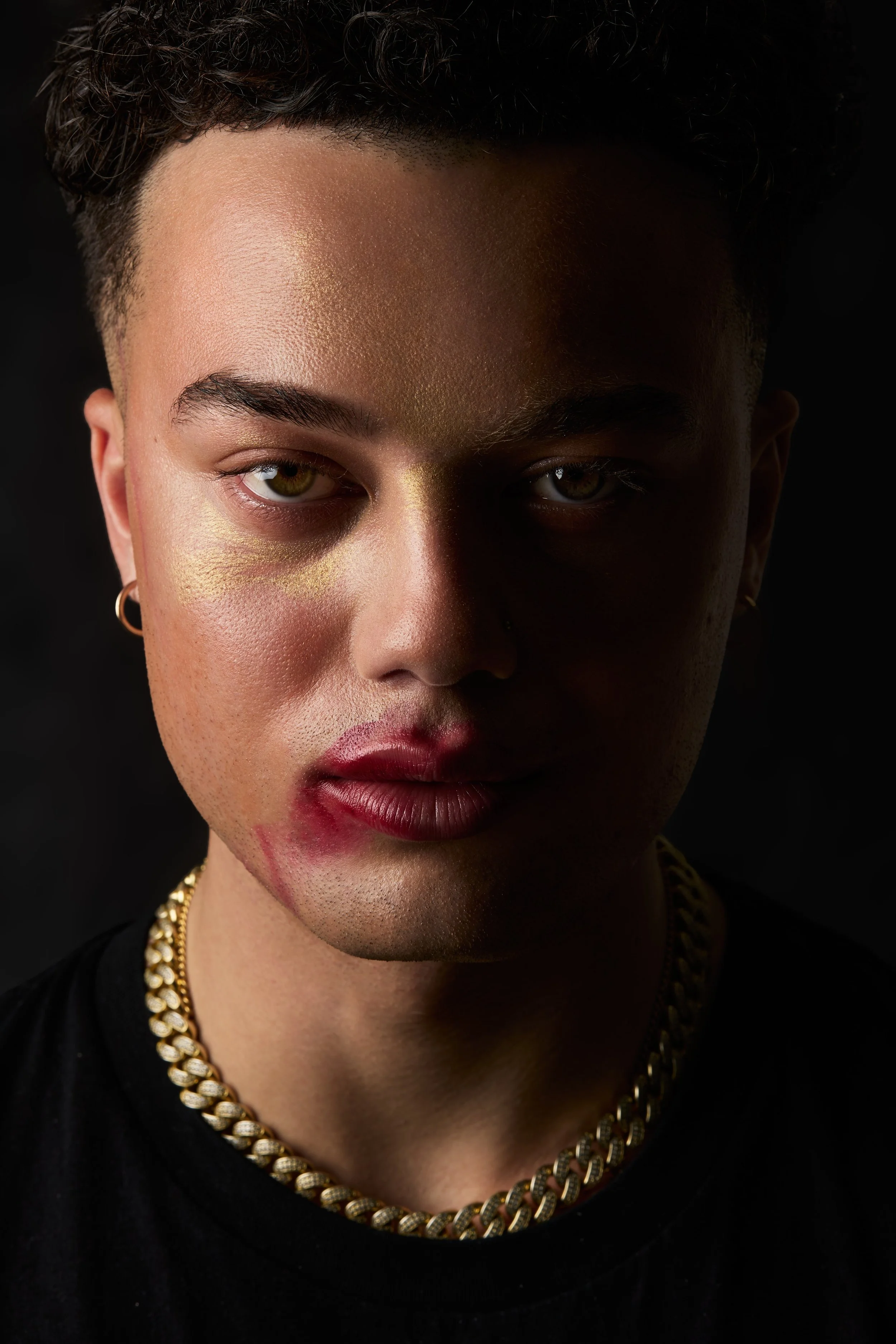 A close-up portrait of a person with short dark hair, gold makeup accents under one eye, gold earrings, layered gold necklaces, and makeup resembling lipstick smudged on their lips, against a dark background.