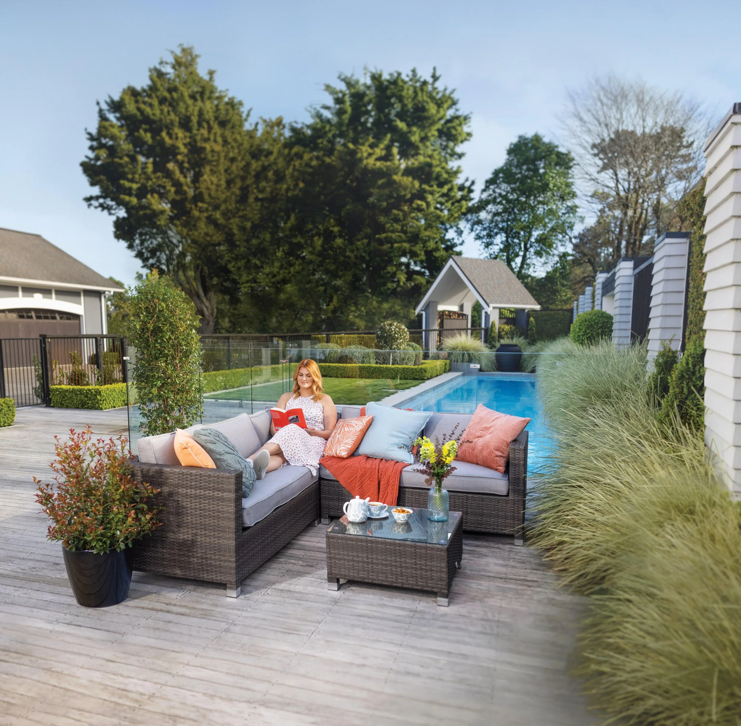 A woman sitting on outdoor patio furniture by a swimming pool, reading a book, with cushions and a vase of flowers on a small table beside her, surrounded by lush greenery and tall trees. An advertisement for a homewares store.