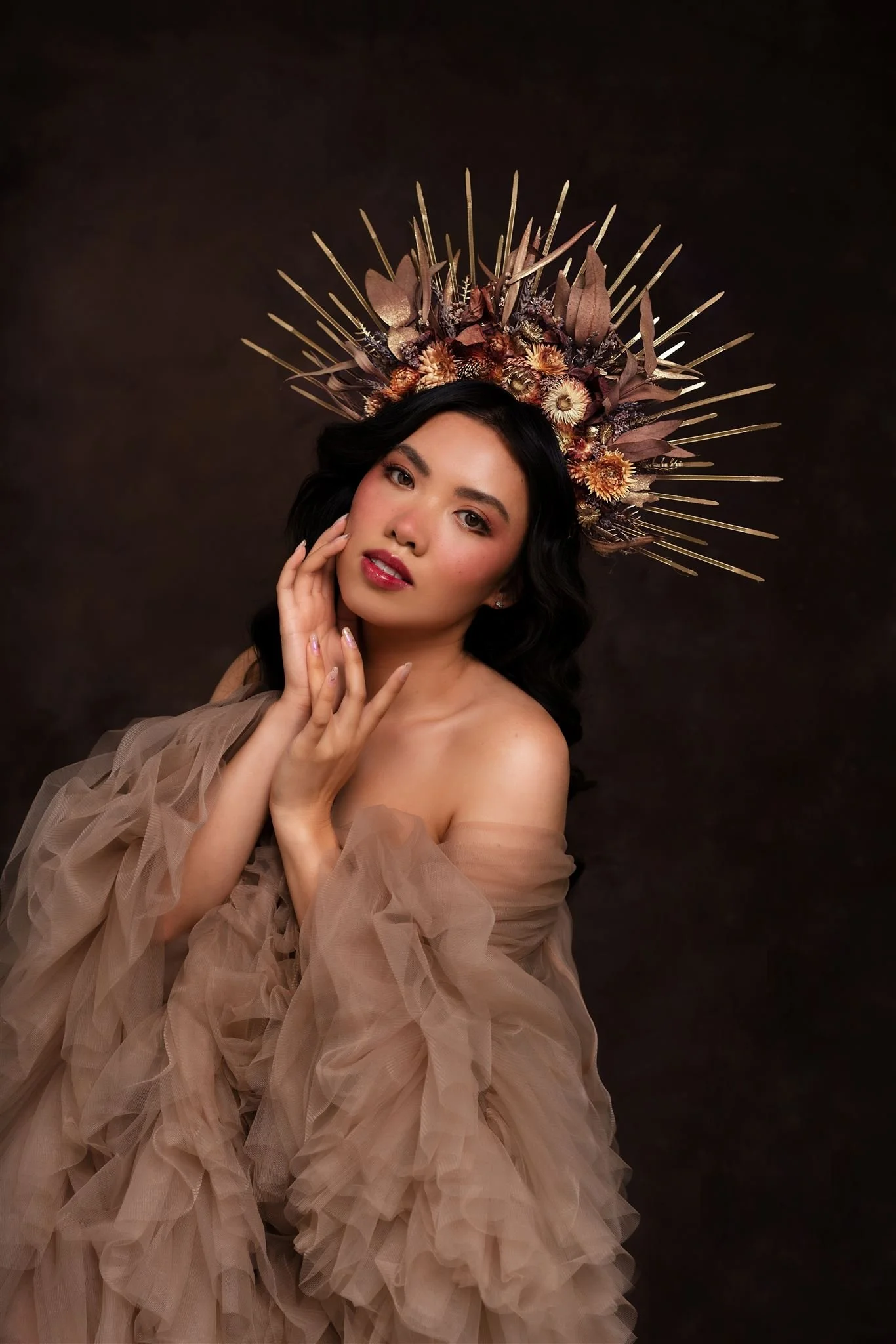 A woman with dark, wavy hair and makeup, wearing an off-shoulder, beige tulle dress, posing with one hand touching her face against a dark background, and wearing a large, ornate headpiece made of dried flowers and geometric spikes.