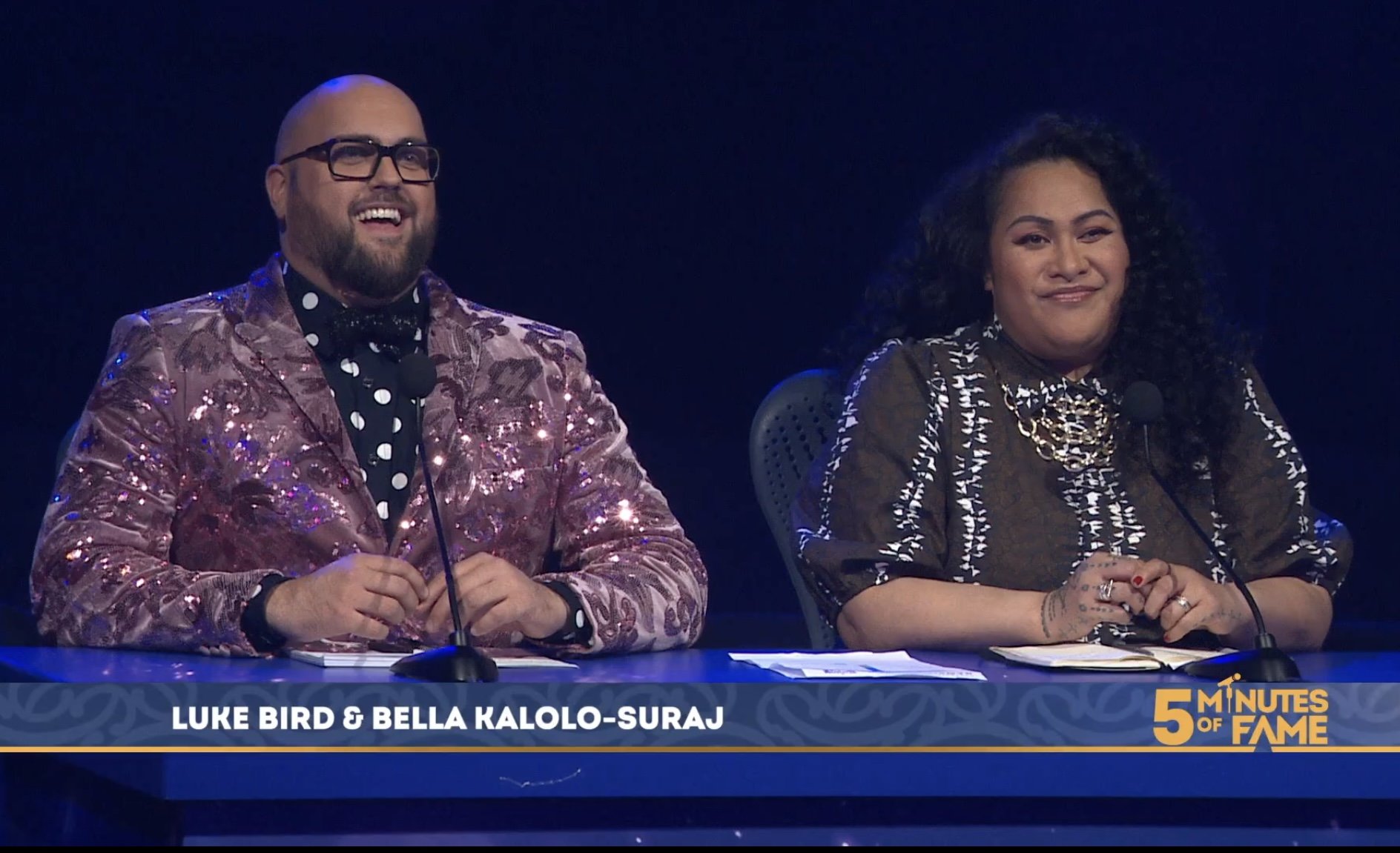 Two judges sitting at a panel, one man dressed in a shiny pink jacket and glasses, one woman with curly hair wearing a patterned top. Both are smiling, with microphones in front of them, on a show called '5 Minutes of Fame'.