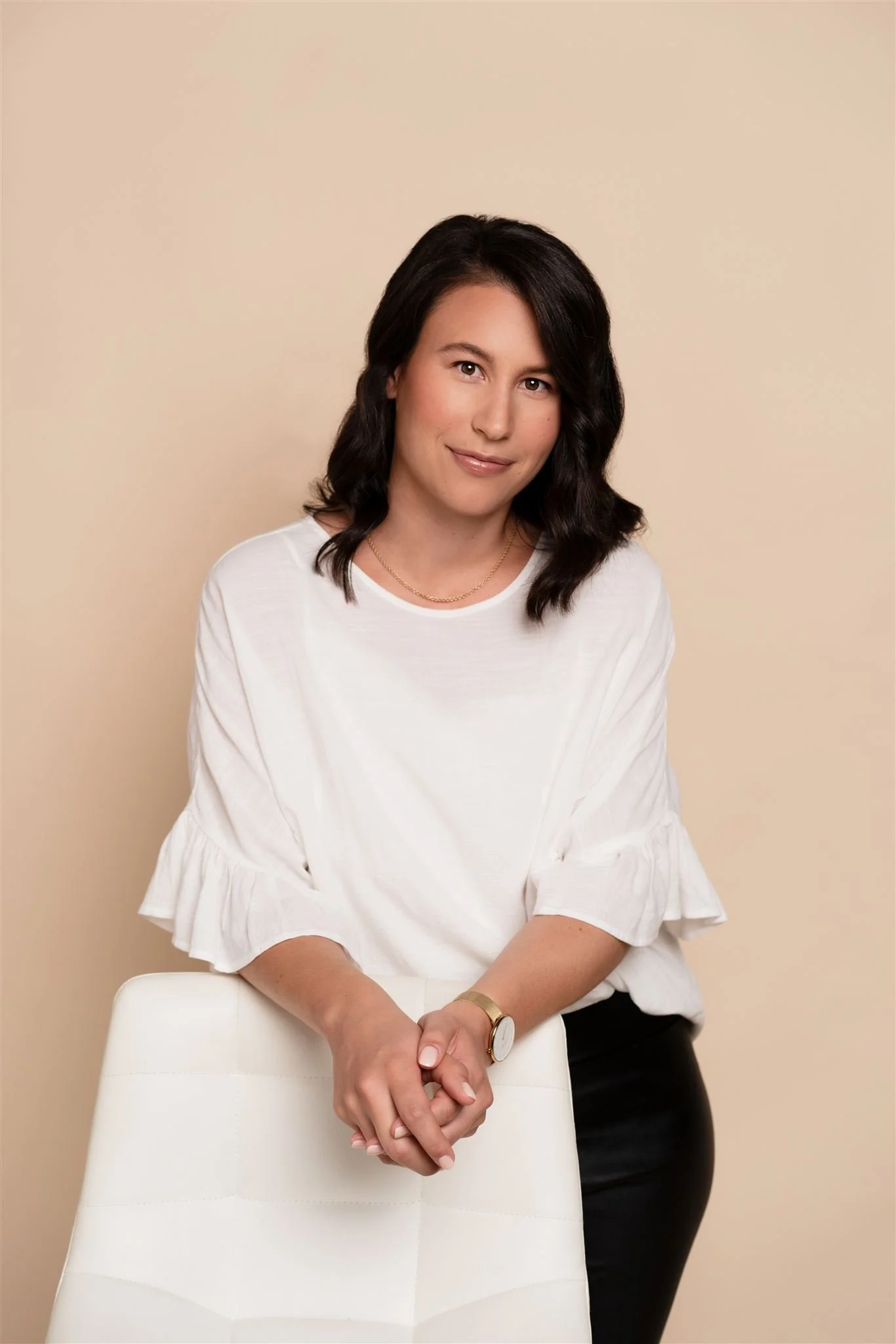 A woman with dark hair, wearing a white blouse with ruffled sleeves, leaning on a white chair against a beige background.