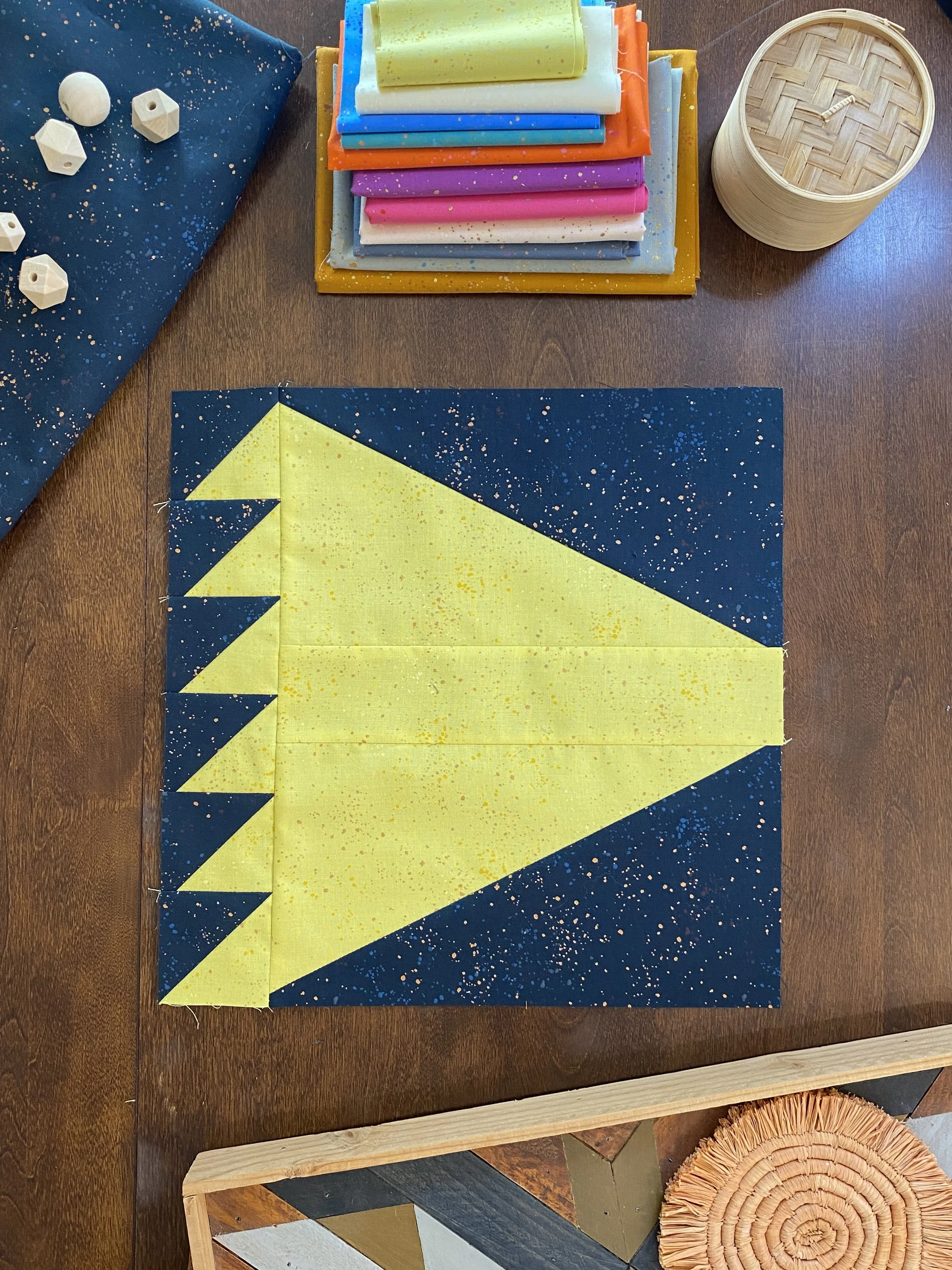 A quilt in progress with a yellow and blue outer space design featuring a large yellow triangle in the center, surrounded by blue fabric with white and yellow speckles, on a wooden table.