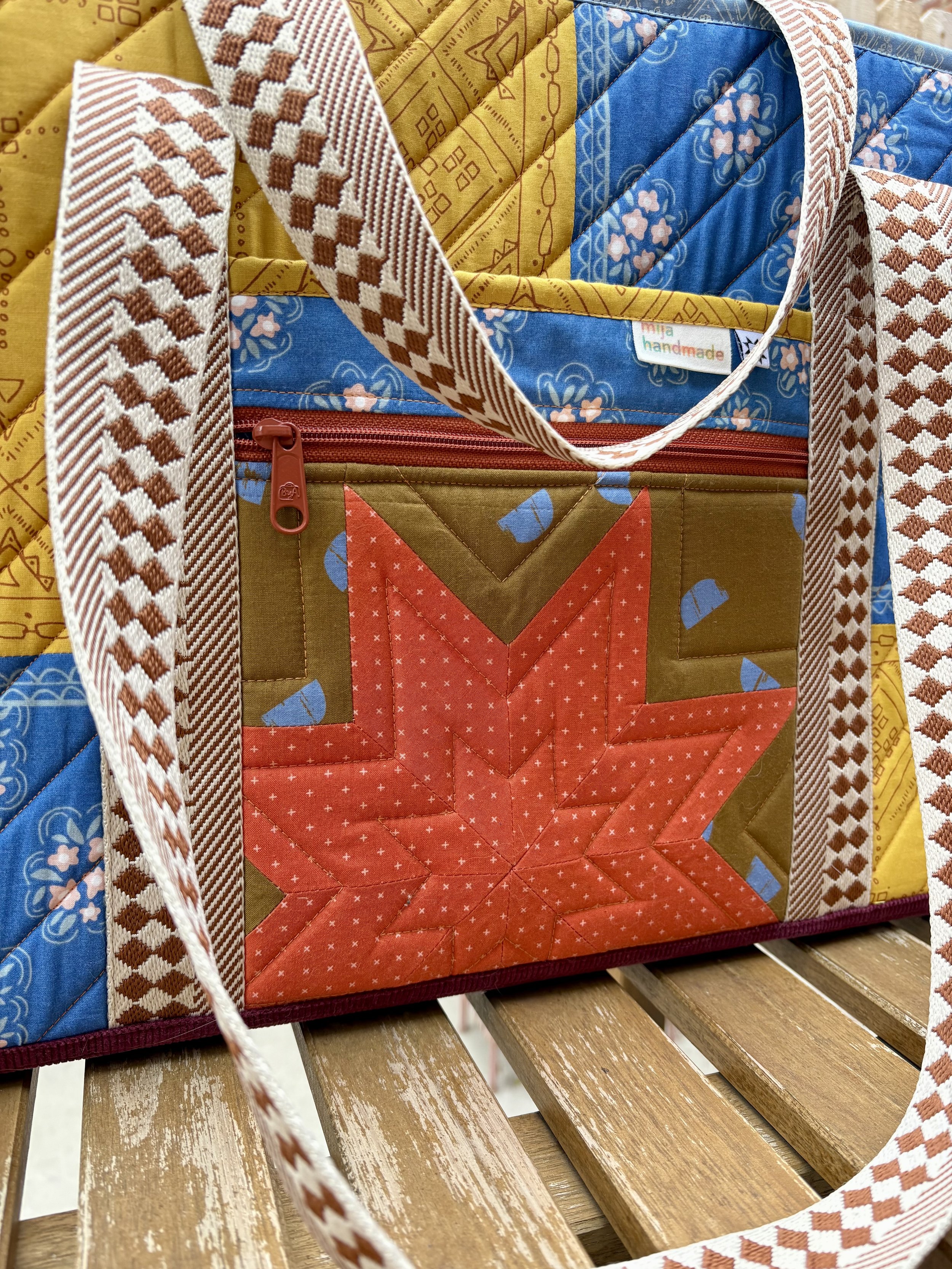 A handmade quilted tote bag featuring blue floral bandana fabric, mustard yellow patchwork, a coral geometric pocket, and a zippered compartment.