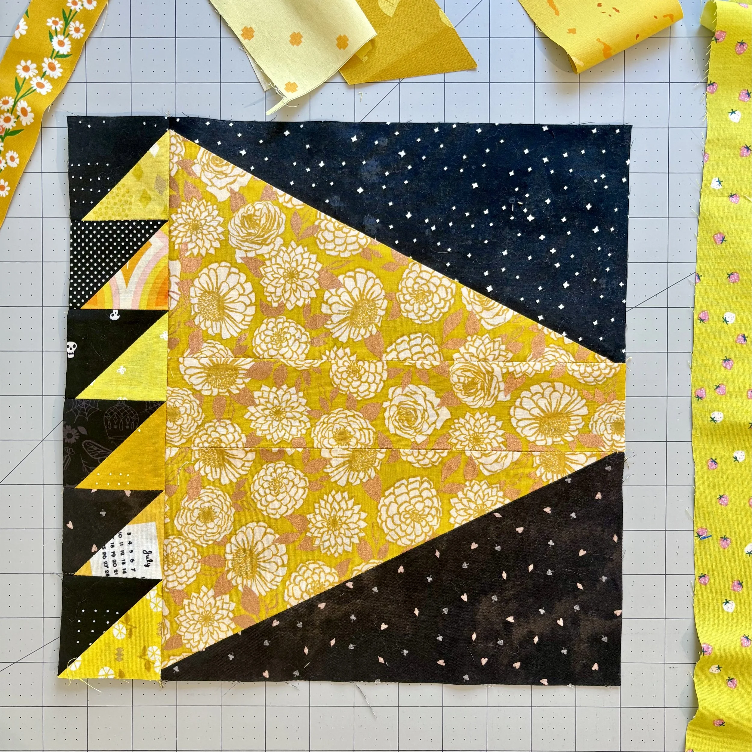 A quilt block featuring a mixture of black, yellow, and floral patterned fabrics arranged in a geometric design, with additional fabric strips on the grid background.