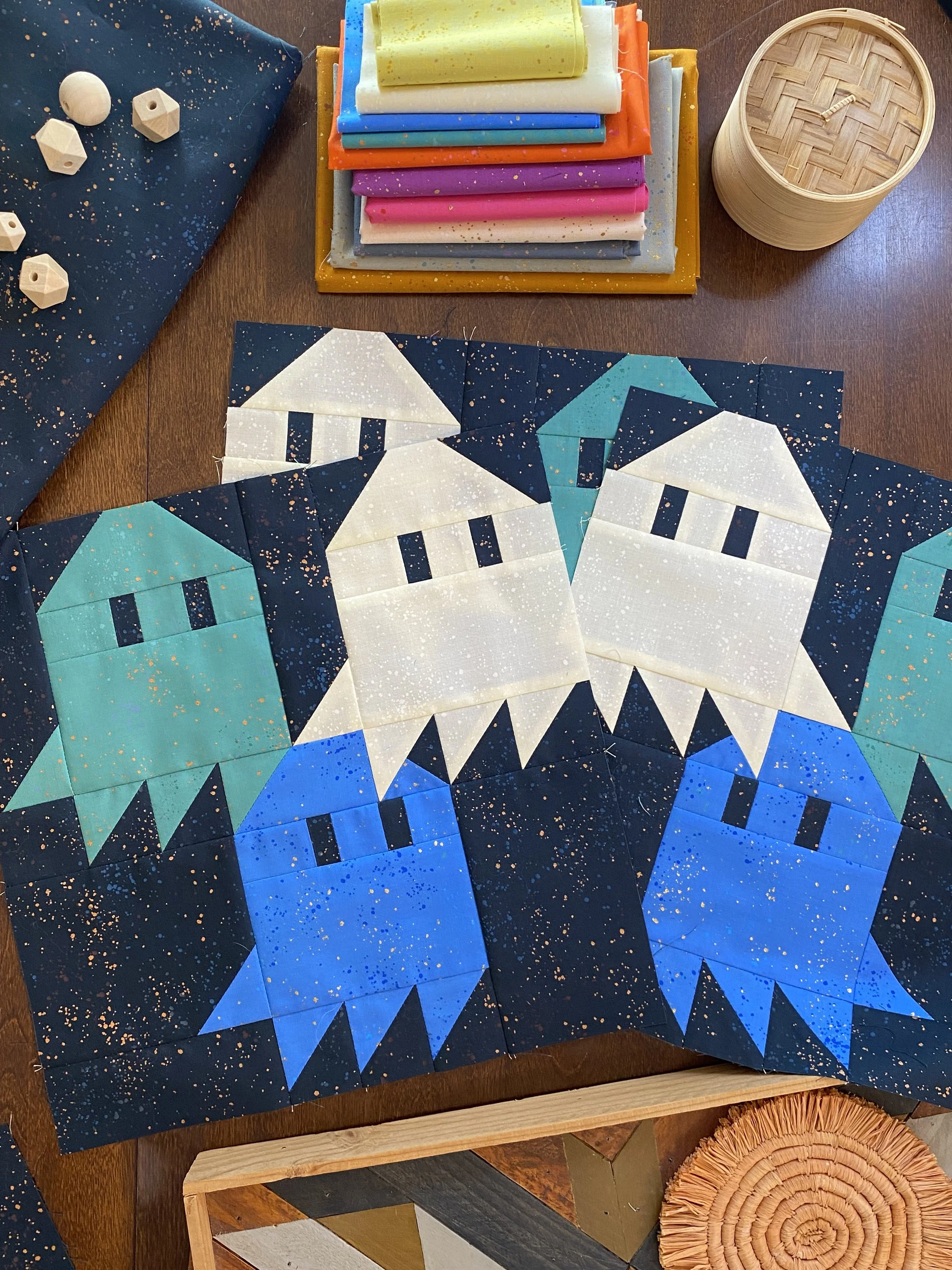 Colorful quilt blocks with a space-themed design featuring white, blue, and teal rocket ships on a dark starry background, arranged on a table with fabric, sewing tools, and other craft supplies around.