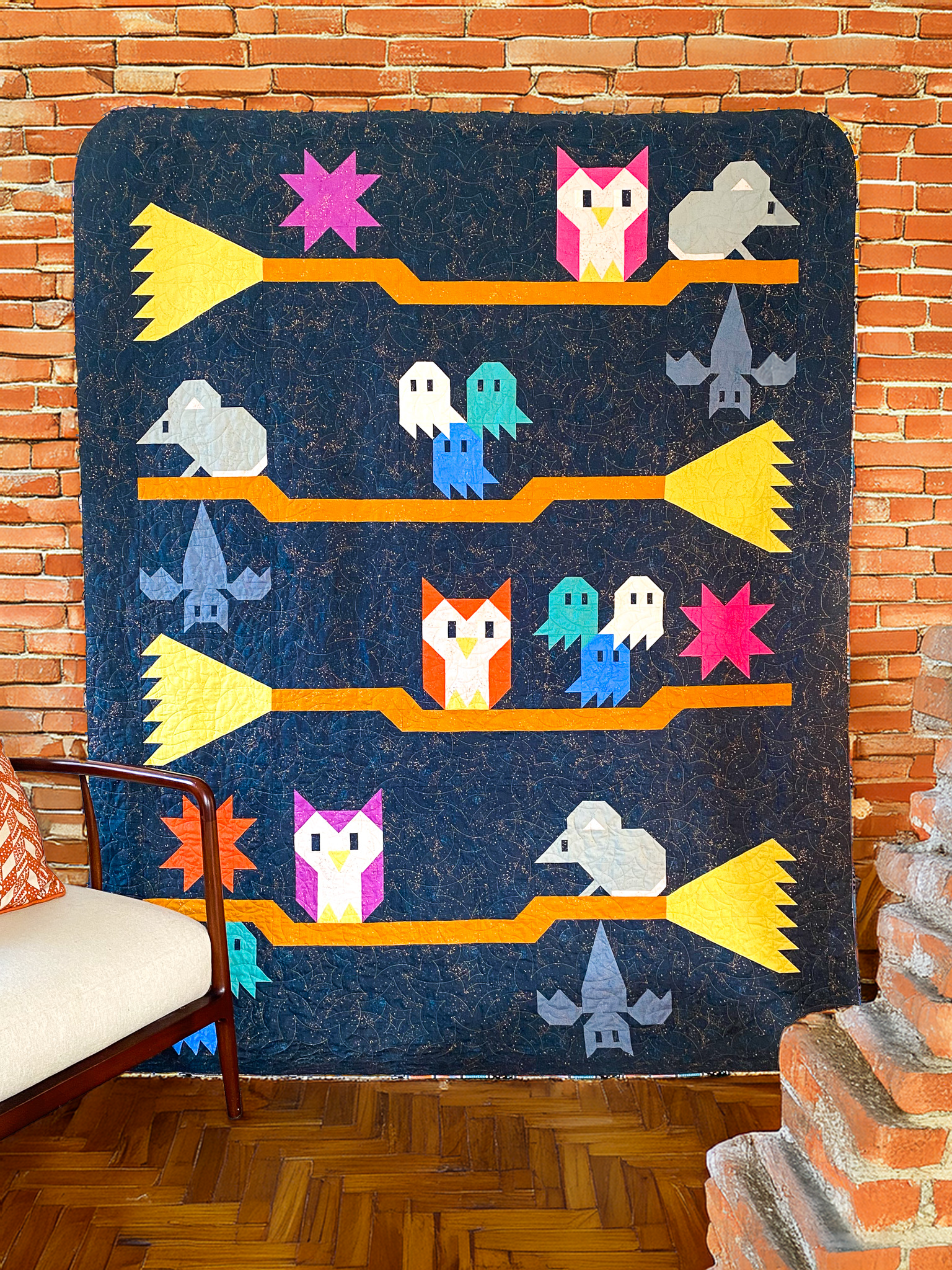 Colorful handmade quilt with pixel art style cats, owls, bats, ghosts, stars, and rockets hanging on a brick wall.