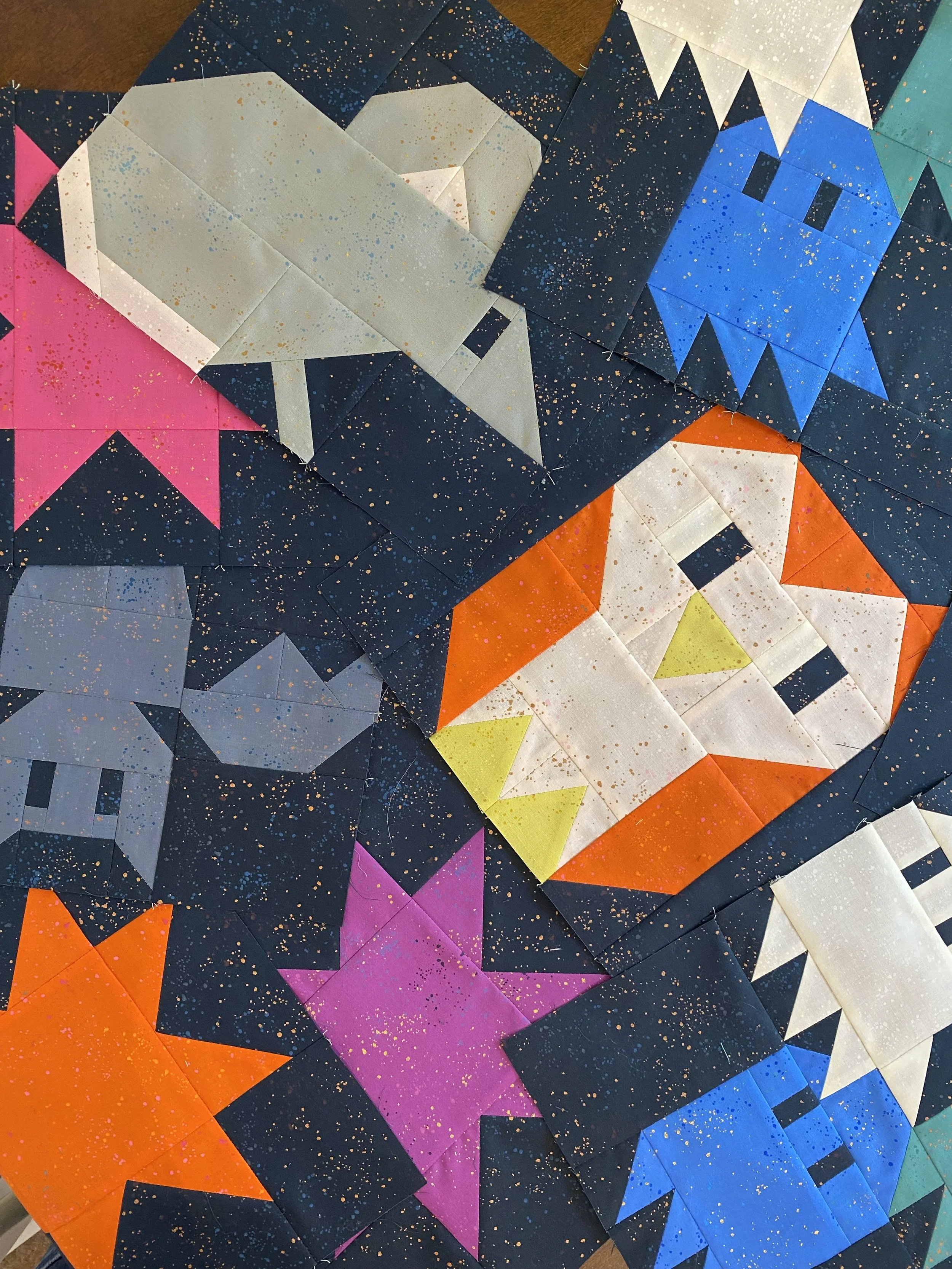 Colorful quilt patches with pixelated Halloween-themed designs including a white ghost, an orange pumpkin with a candle, a gray skull, a purple bat, a blue Frankenstein face, and a white witch's hat, all on a dark background splattered with gold paint.