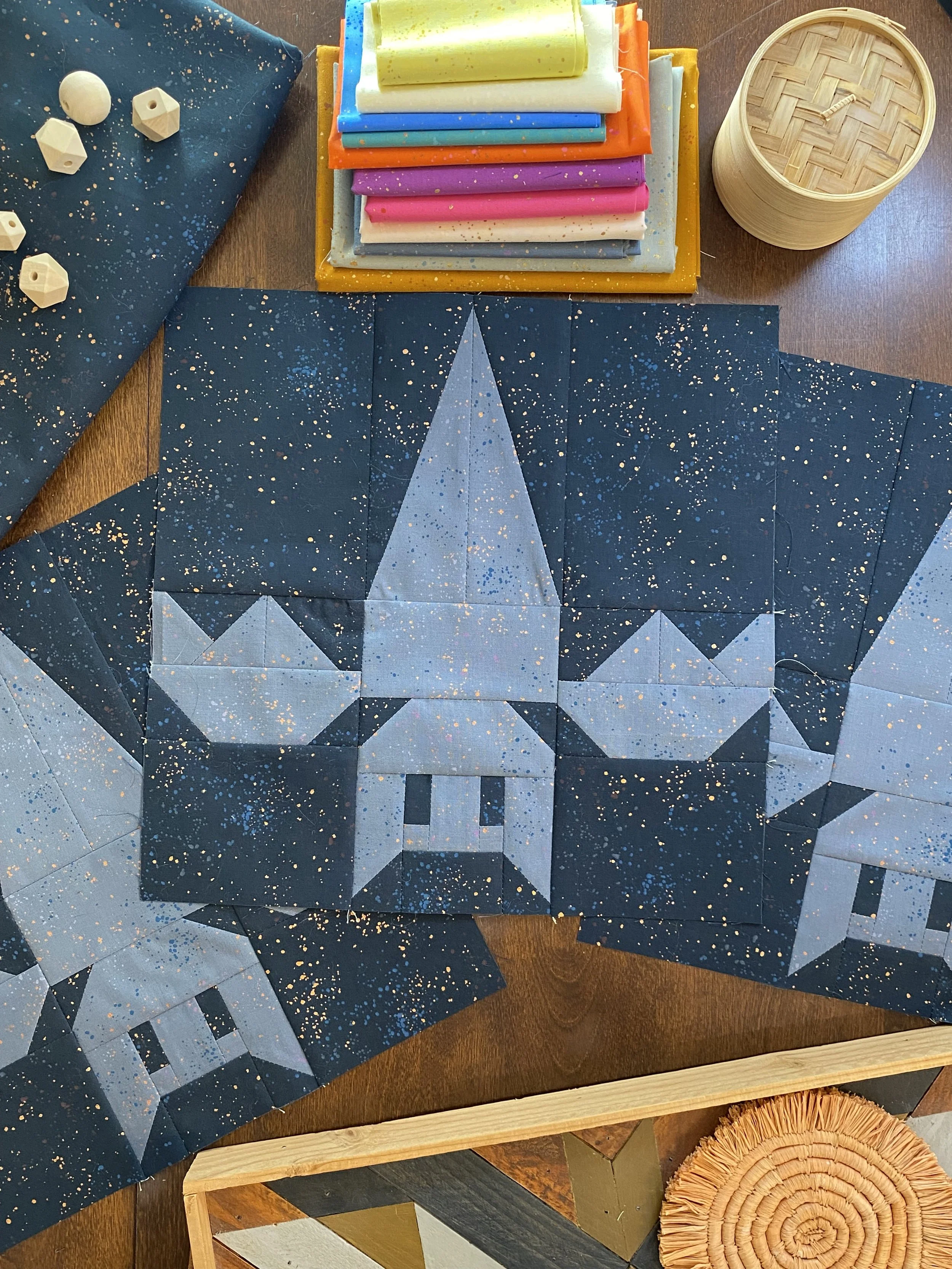 A quilting project in progress features a space-themed star design with a character resembling a wizard or mage on dark blue fabric with white speckles, surrounded by sewing supplies like colorful fabric, white plastic sewing machine parts, and a wooden basket.