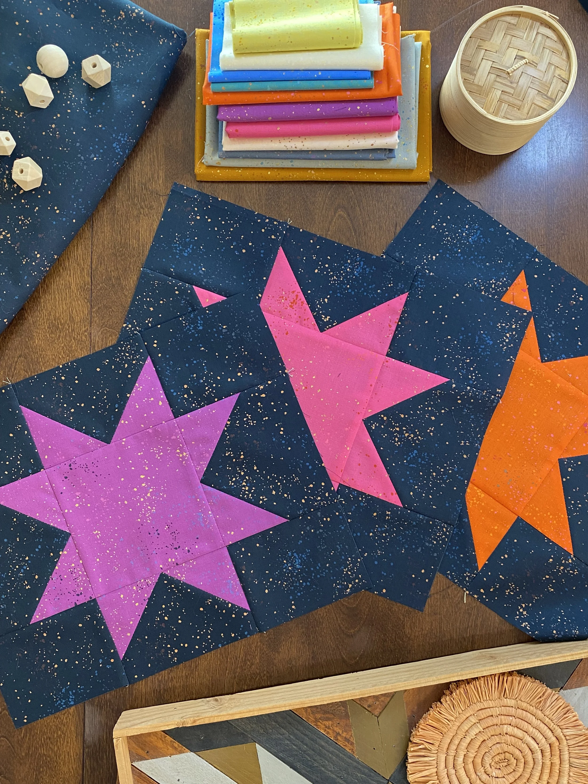 Colorful quilt blocks with star patterns on a wooden table. The quilt blocks are dark blue with pink, purple, and orange stars, all speckled with paint or ink. Surrounding items include fabric rolls in various colors, a round bamboo container, and a stack of white wooden beads.