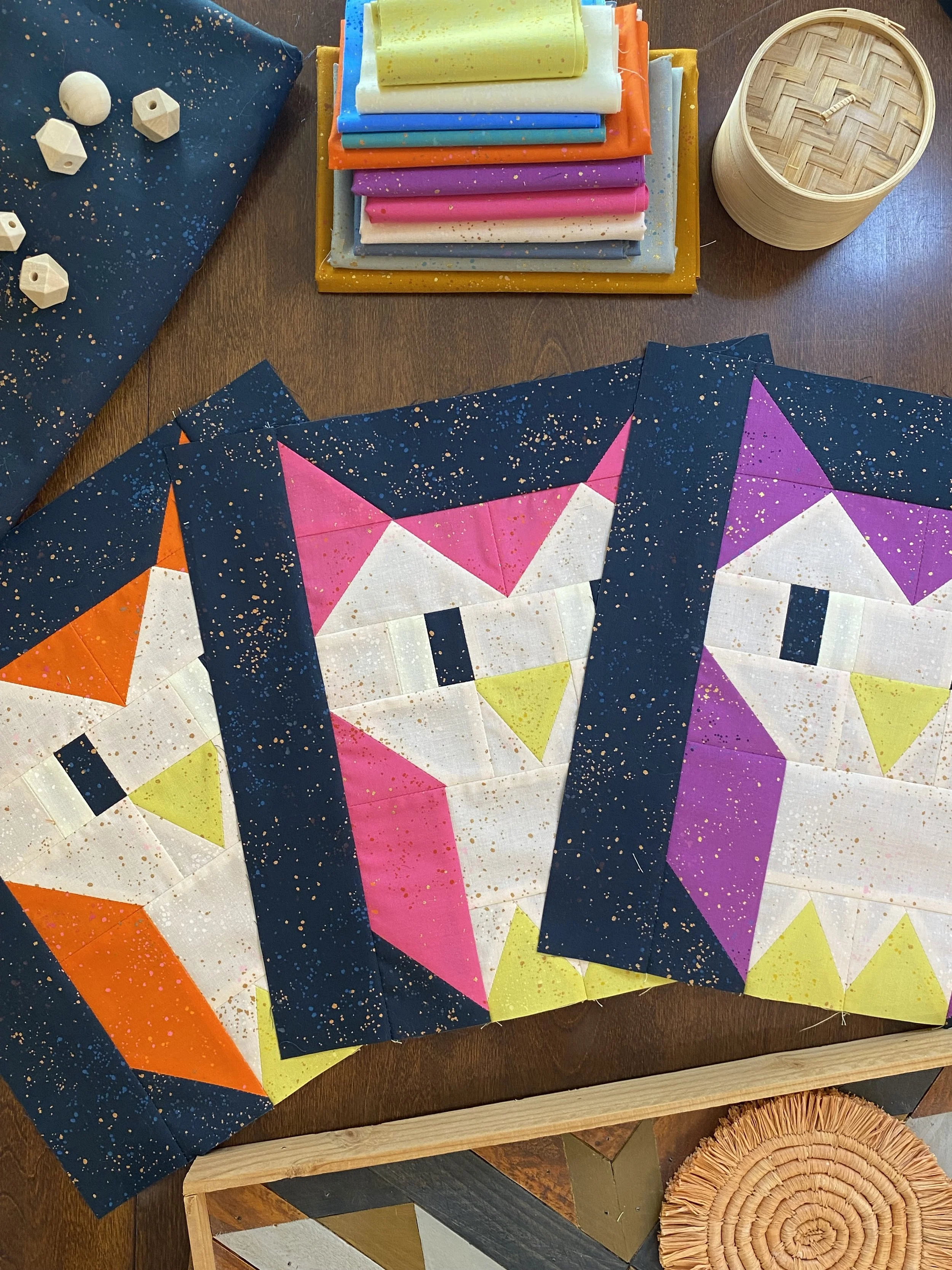 Three colorful quilt blocks with geometric designs of cats, in pink, purple, and orange shades, laid out on a wooden surface.